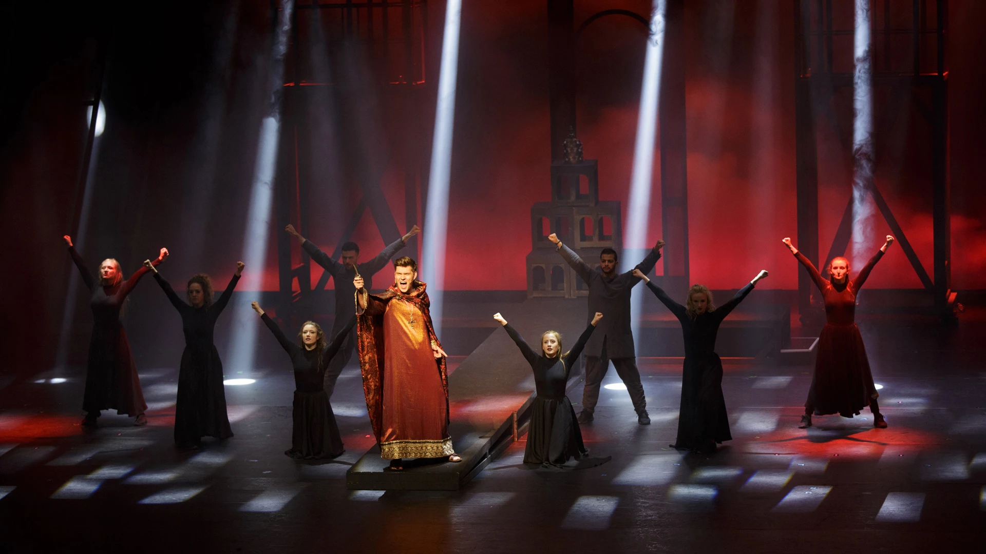 Im dämmrigen Szenenmotiv im die Päpstin Musical ist ein Sänger im Rot- Braun-Goldenem Gewand dargestellt, umgeben von weiteren Darstellern