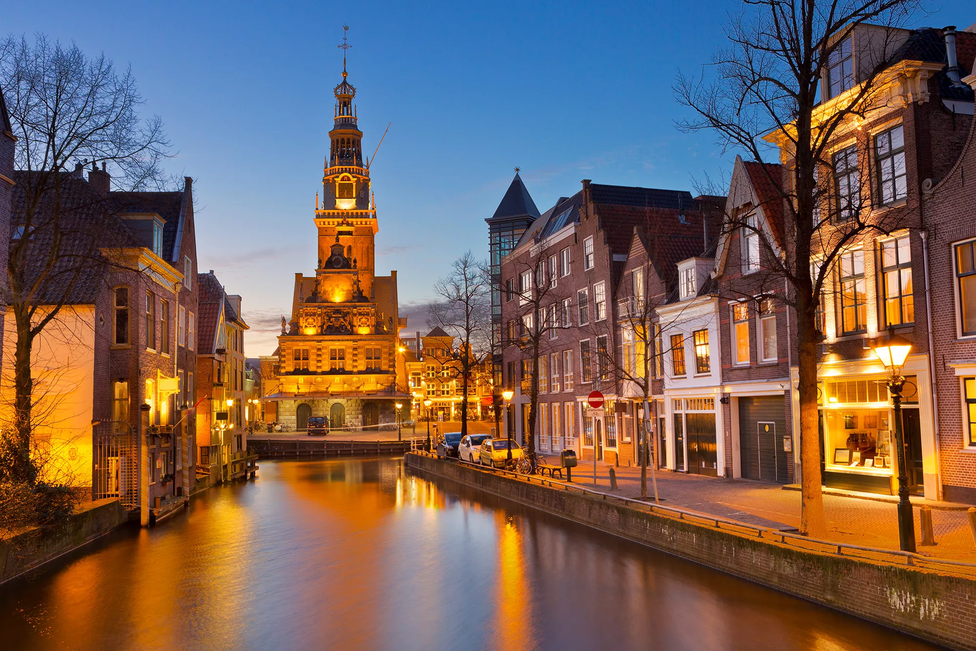 Abendansicht des Nordhollandkanals in Alkmaar, erleuchtet von städtischer Beleuchtung. Das warme Licht reflektiert sanft auf der Wasseroberfläche und sorgt für eine malerische Atmosphäre. Umgeben von historischen Gebäuden und sanften Wellen bietet die Szene eine einladende und romantische Stimmung.