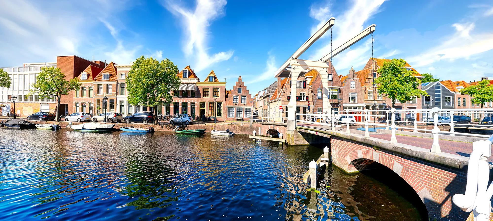 Blick auf die Brücke über den Oudegracht-Kanal in Alkmaar bei Tageslicht, umgeben von malerischen Gebäuden. Am Ufer sind mehrere angelegte Boote zu sehen, die eine charmante maritime Atmosphäre schaffen. Die Sonne reflektiert auf dem Wasser und verleiht der Szene einen einladenden Glanz.