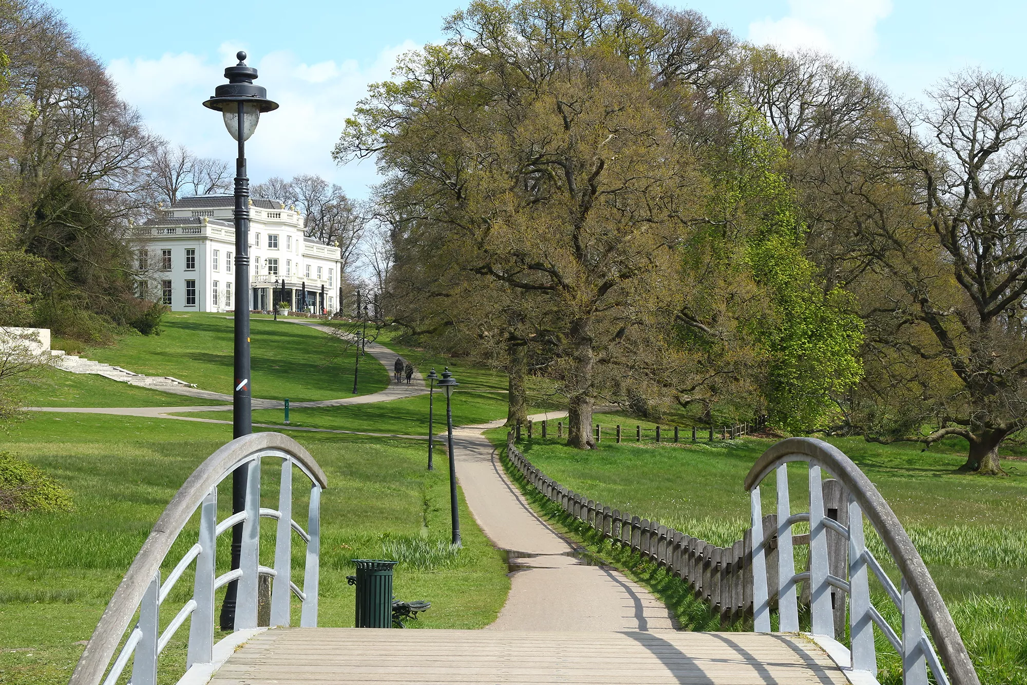 Ein Fußweg führt über die Brücke im Park Sonsbeek zu einer eleganten weißen Villa, umgeben von üppigen grünen Wiesen und schattenspendenden Bäumen, die eine friedliche und einladende Atmosphäre schaffen.