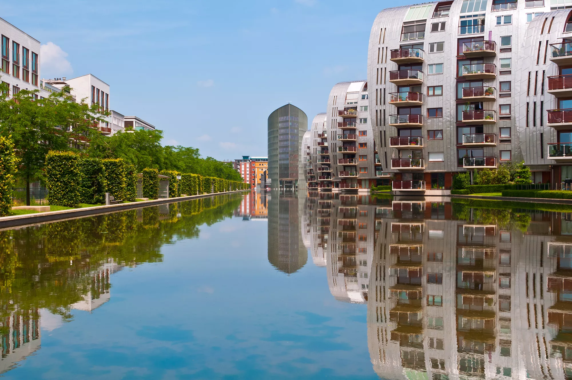 Die Stadt Hertogenbosch präsentiert sich im malerischen Stadtteil Paleiskwartier unter strahlendem Sonnenschein und klarem Himmel. Die modernen Architektur und historischen Gebäude harmonieren perfekt mit der grünen Umgebung, während die lebhaften Farben der Fassaden und die blühenden Pflanzen eine einladende Atmosphäre schaffen. Die Umgebung spiegelt sich malerisch im Den Bosch Kanal wider, was dem Anblick eine zusätzliche Dimension verleiht und die idyllische Stimmung der Stadt unterstreicht. Ein idealer Ort, um die Schönheit von Hertogenbosch zu genießen und ihre kulturelle Vielfalt zu erleben.