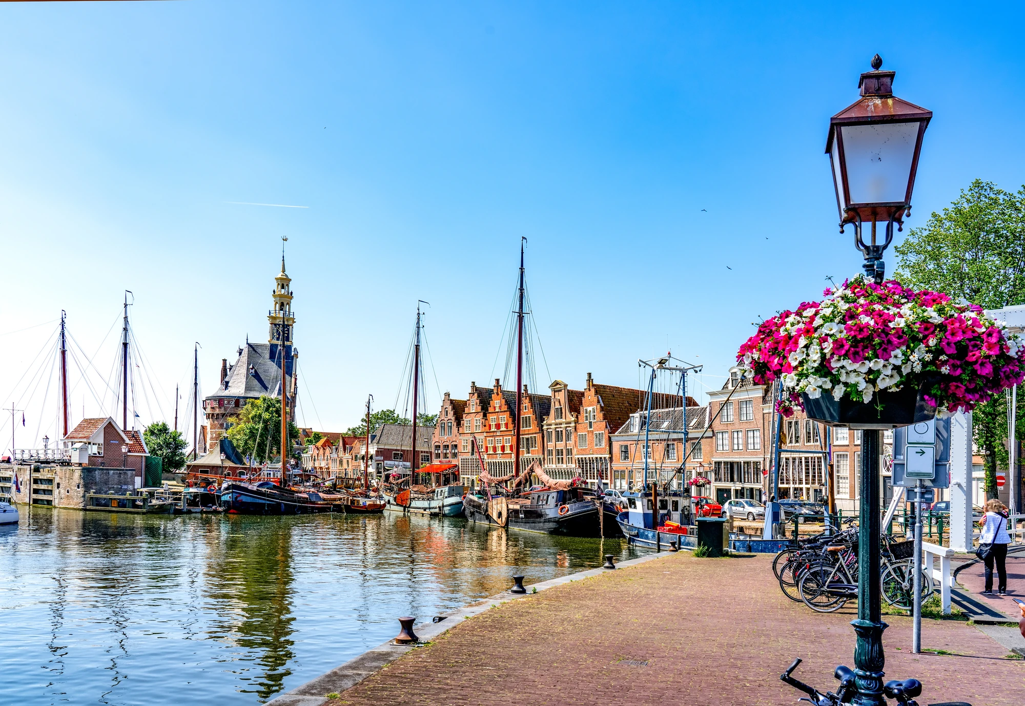 Der historische Hafen in Hoorn, bei Sonnenschein, mit malerischen Häusern und angelegten Segelschiffen. Dekorierte Straßenlaternen, geschmückt mit farbenfrohen Blumen, verleihen der Szene eine einladende und lebendige Atmosphäre. Diese Kombination aus maritimer Geschichte und blühendem Stadtleben macht den Hafen zu einem charmanten Anziehungspunkt.