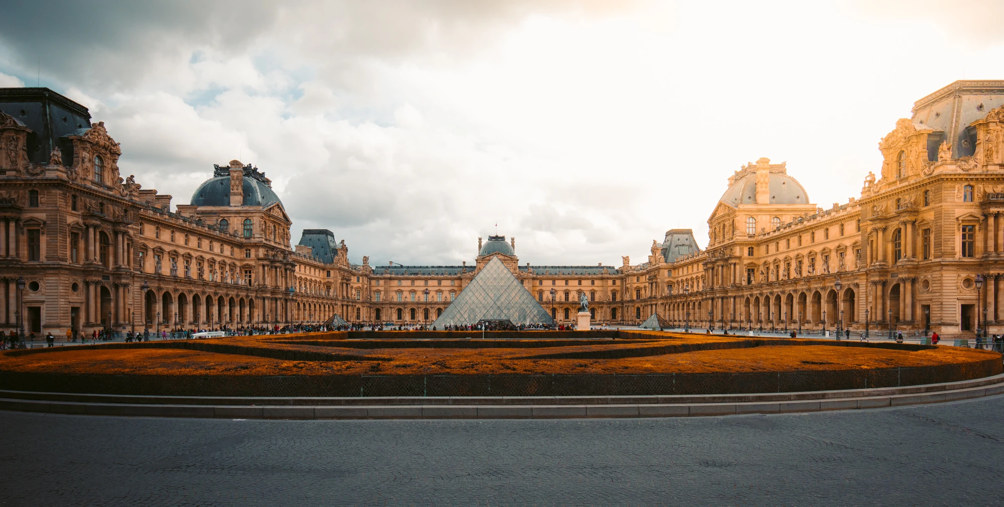 Außenansicht des Louvre in Paris bei leicht bewölkter Abenddämmerung, mit sanften Lichtverhältnissen, die die beeindruckende Architektur betonen.