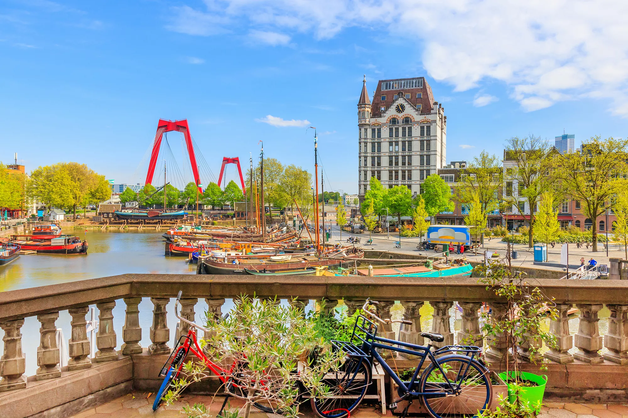 Blick auf den Oude Haven in Rotterdam, mit historischen Booten im Wasser und der ikonischen roten Willemsbrücke im Hintergrund. Die Szenerie verbindet das maritime Erbe des Hafens mit moderner Infrastruktur, während die lebhafte Atmosphäre und der Kontrast zwischen Alt und Neu die Vielseitigkeit der Stadt widerspiegeln.