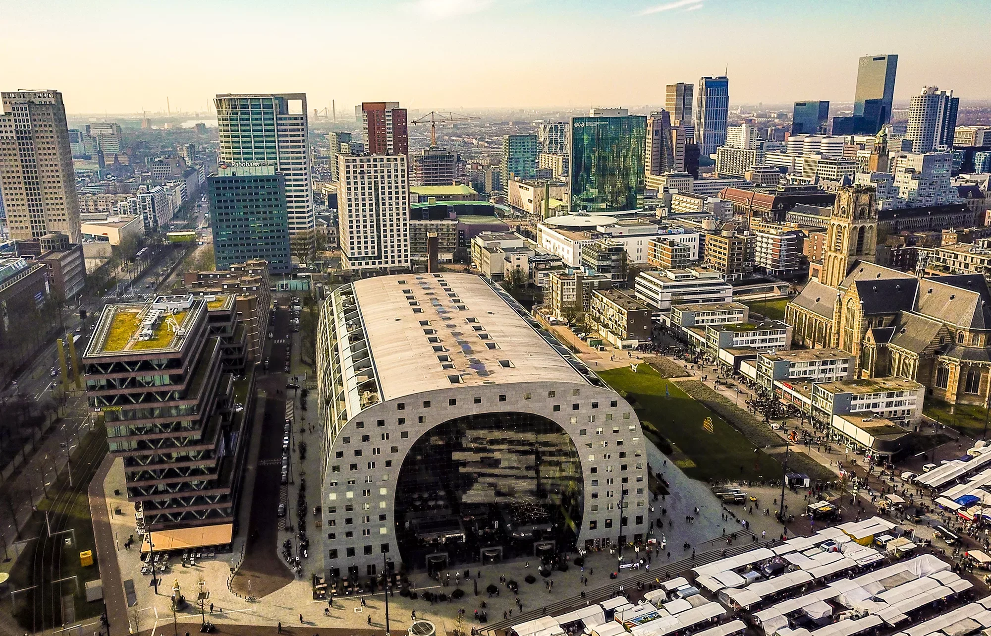 Ein beeindruckender Blick von oben auf die Markthalle und das Zentrum von Rotterdam. Die moderne Architektur der Markthalle sticht hervor, umgeben von den lebendigen Straßen und den umliegenden Gebäuden des Stadtzentrums. Die harmonische Mischung aus urbanem Leben und architektonischer Innovation verleiht der Szenerie eine dynamische und zeitgenössische Atmosphäre.