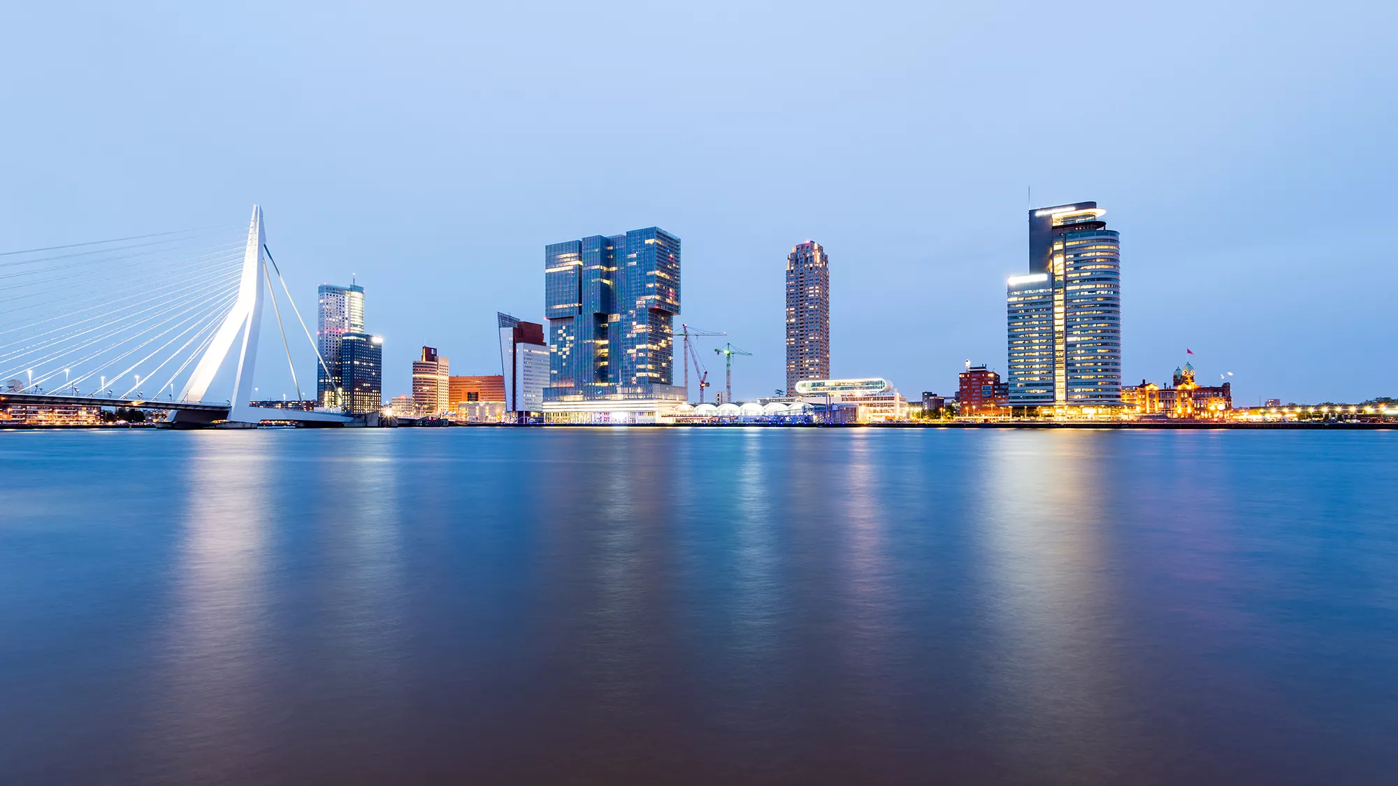 Ein Blick auf die Erasmusbrücke und die beleuchtete Skyline von Rotterdam am Abend. Die Lichter der Brücke und der Gebäude reflektieren sich malerisch auf der Wasseroberfläche, wodurch eine stimmungsvolle und lebendige Szenerie entsteht. Die ikonische Brücke und die moderne Architektur fangen den urbanen Charme Rotterdams in der nächtlichen Atmosphäre ein.