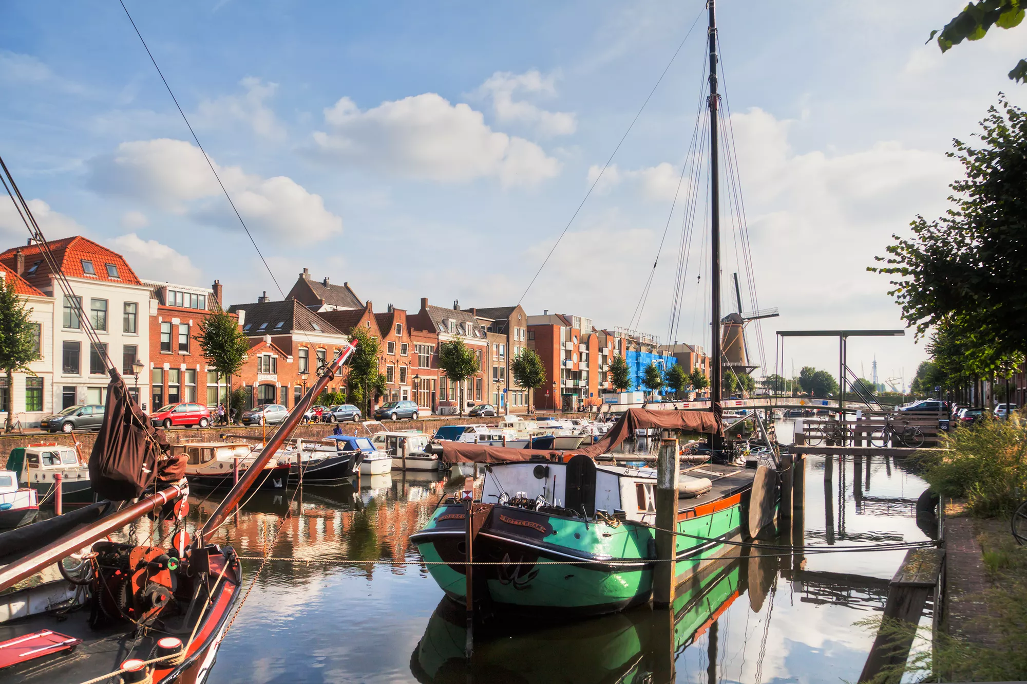 Historischer Delfshaven in Rotterdam, mit angelegten Booten entlang des ruhigen Wassers. Dahinter erstreckt sich eine charmante Häuserreihe, vor der geparkte Autos zu sehen sind. Einige Menschen spazieren entspannt entlang der Promenade und genießen die malerische Atmosphäre. Diese Szenerie verbindet die maritime Geschichte mit dem lebendigen Stadtleben.