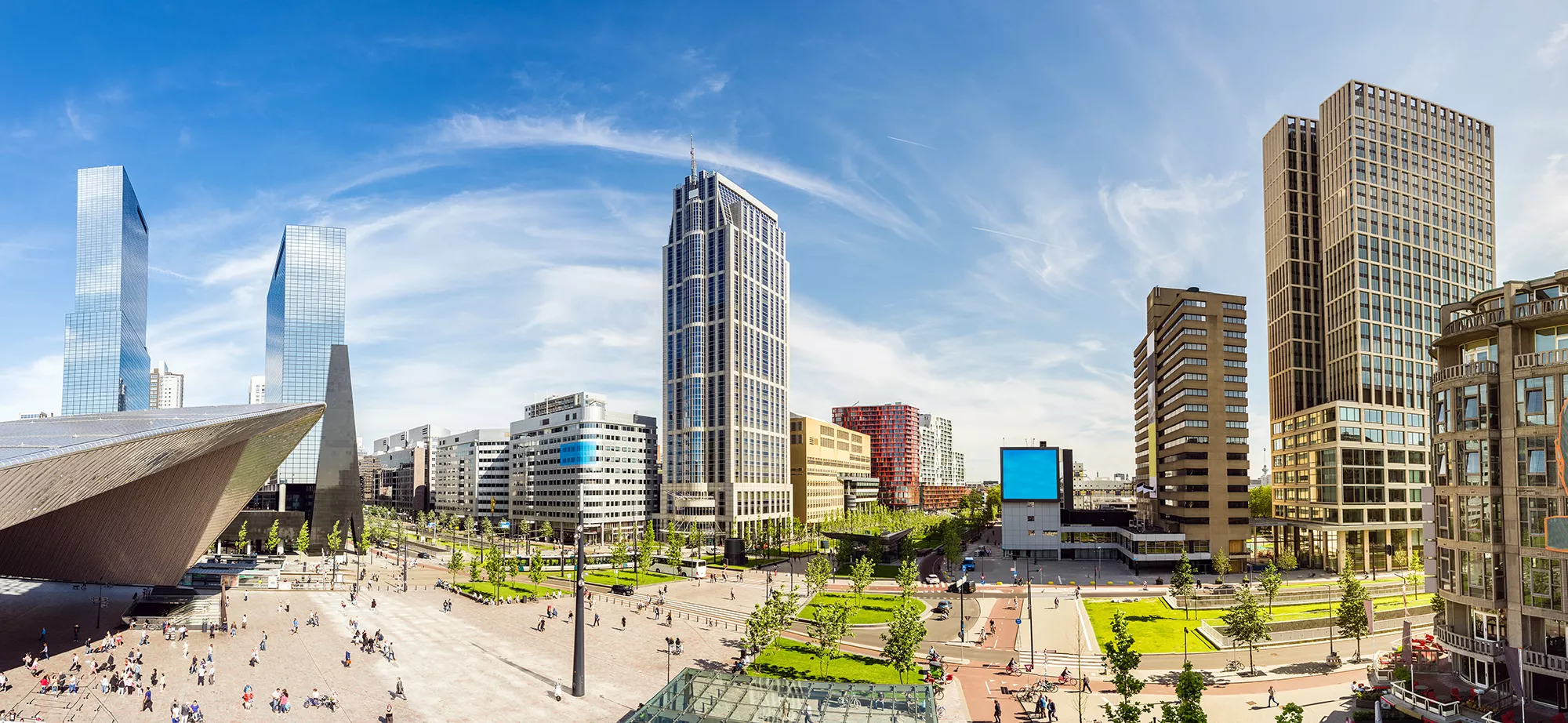 Blick auf den Bahnhof und die Skyline von Rotterdam an einem sonnigen Tag. Die strahlende Sonne hebt die moderne Architektur hervor, während Menschen unten entlang der Straßen und Gehwege gehen. Die lebhafte Atmosphäre der Stadt, kombiniert mit der imposanten Skyline, vermittelt ein Bild von urbaner Dynamik und Energie.
