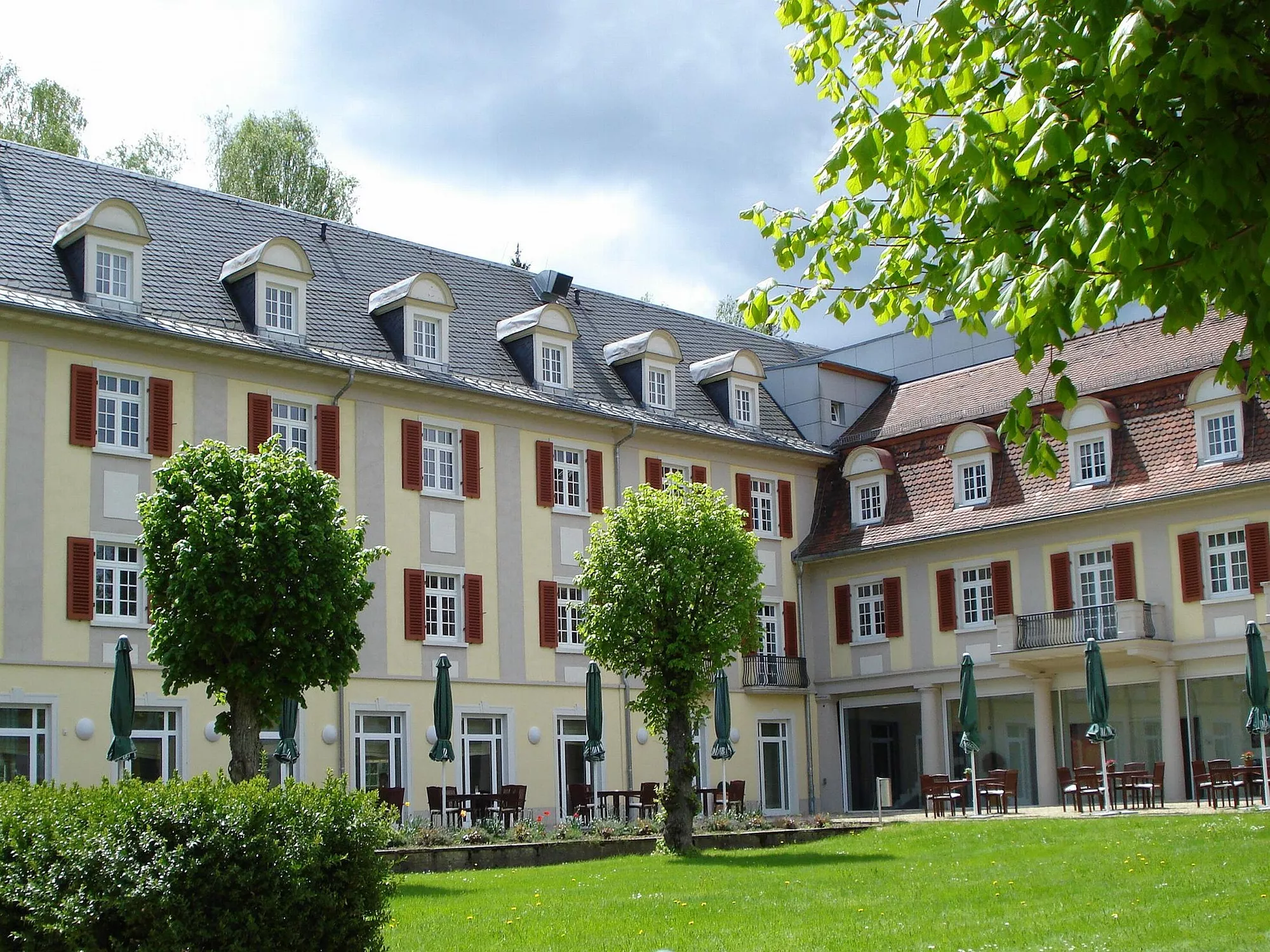 Rückansicht des Santé Royale Bad Brambach mit in zarten Farbtönen gehaltener Fassade und davor stehenden Sitzgruppen mit Sonnenschirm im Grünen