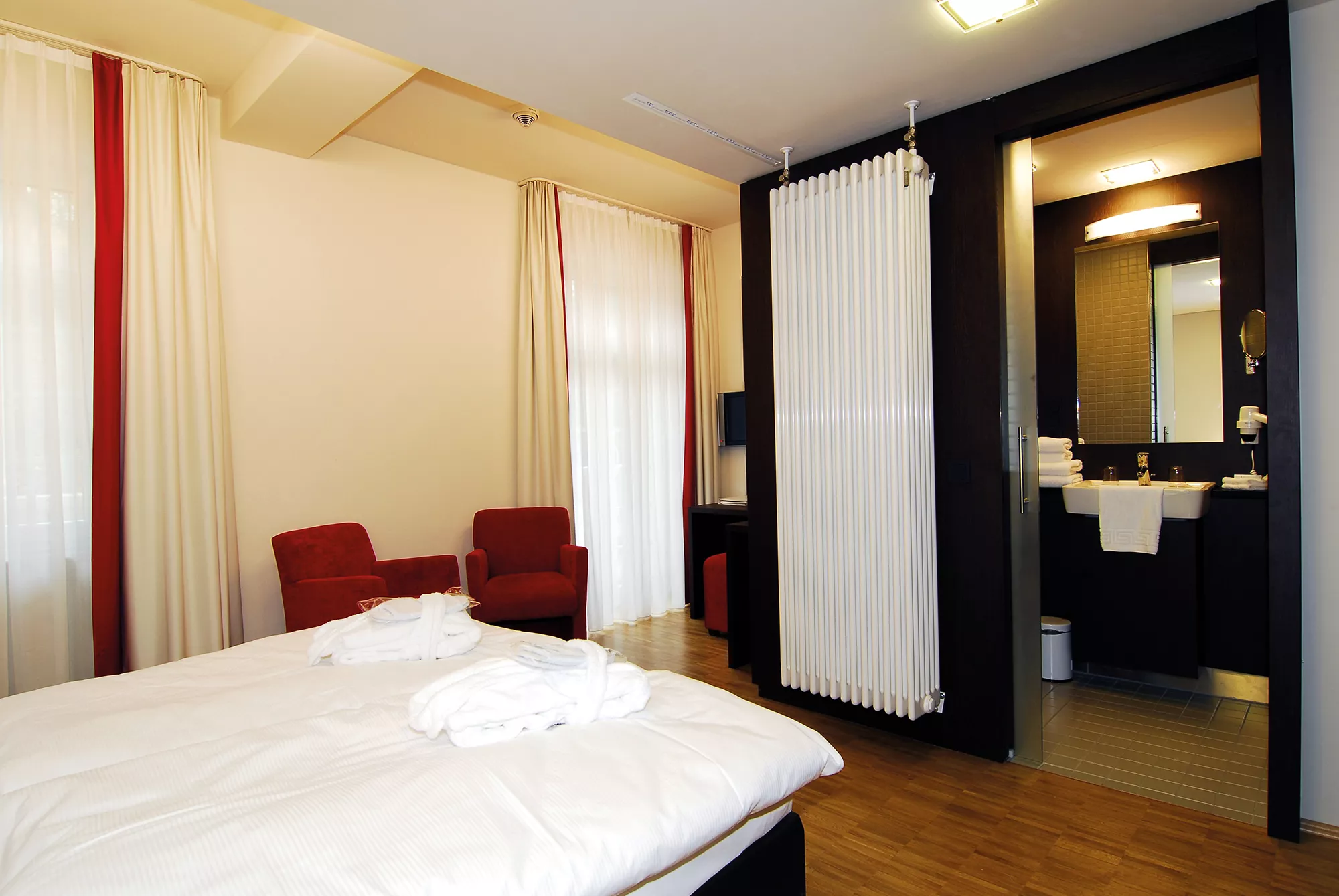 Blick in ein Gästezimmerbeispiel im Santé Royale Bad Brambach mit in zarten Gelb-,Rot- und braunen Farbtönen abgestimmten Mobiliar und Wandfarben