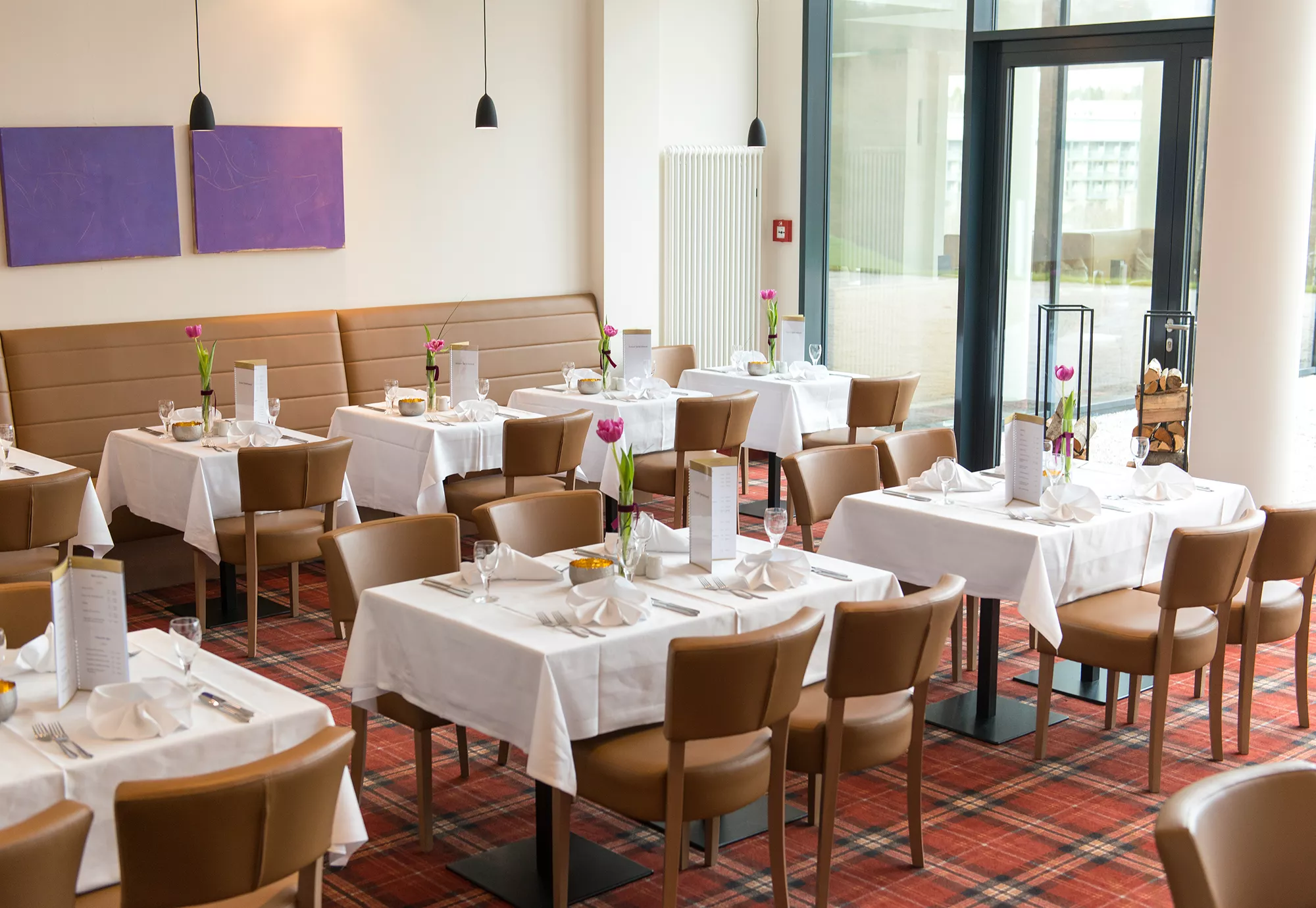 Das Restaurant im Santé Royale Wolkenstein ist in braunen und roten Naturtönen gehalten und mit verschieden großen eingedeckten Tischen dargestellt