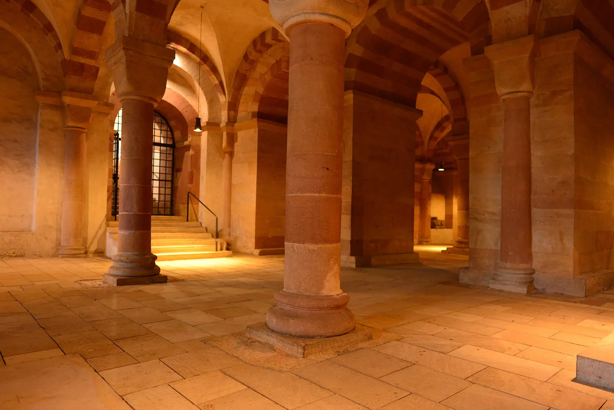 Blick in die beleuchtete Krypta im Dom zu Speyer mit steinernen Säulen und einer nach oben führenden Treppe