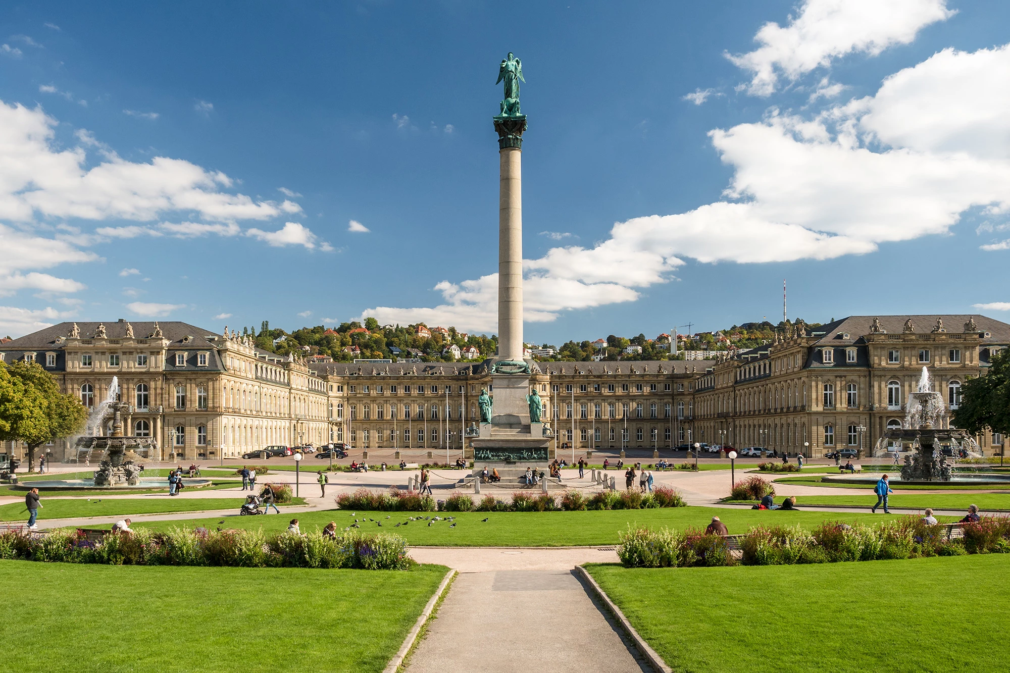 Das Bild zeigt die imposante Jubiläumssäule auf dem Schlossplatz in Stuttgart, umgeben von der prächtigen Architektur des Neuen Schlosses. Die Säule, gekrönt von der Göttin Concordia, ist ein markantes Wahrzeichen und steht inmitten gepflegter Grünanlagen. Der Platz wird von Menschen belebt, die die Sonne genießen, während der Springbrunnen im Vordergrund für eine idyllische Atmosphäre sorgen. Im Hintergrund erstrecken sich die grünen Hügel der Stadt unter einem strahlend blauen Himmel.