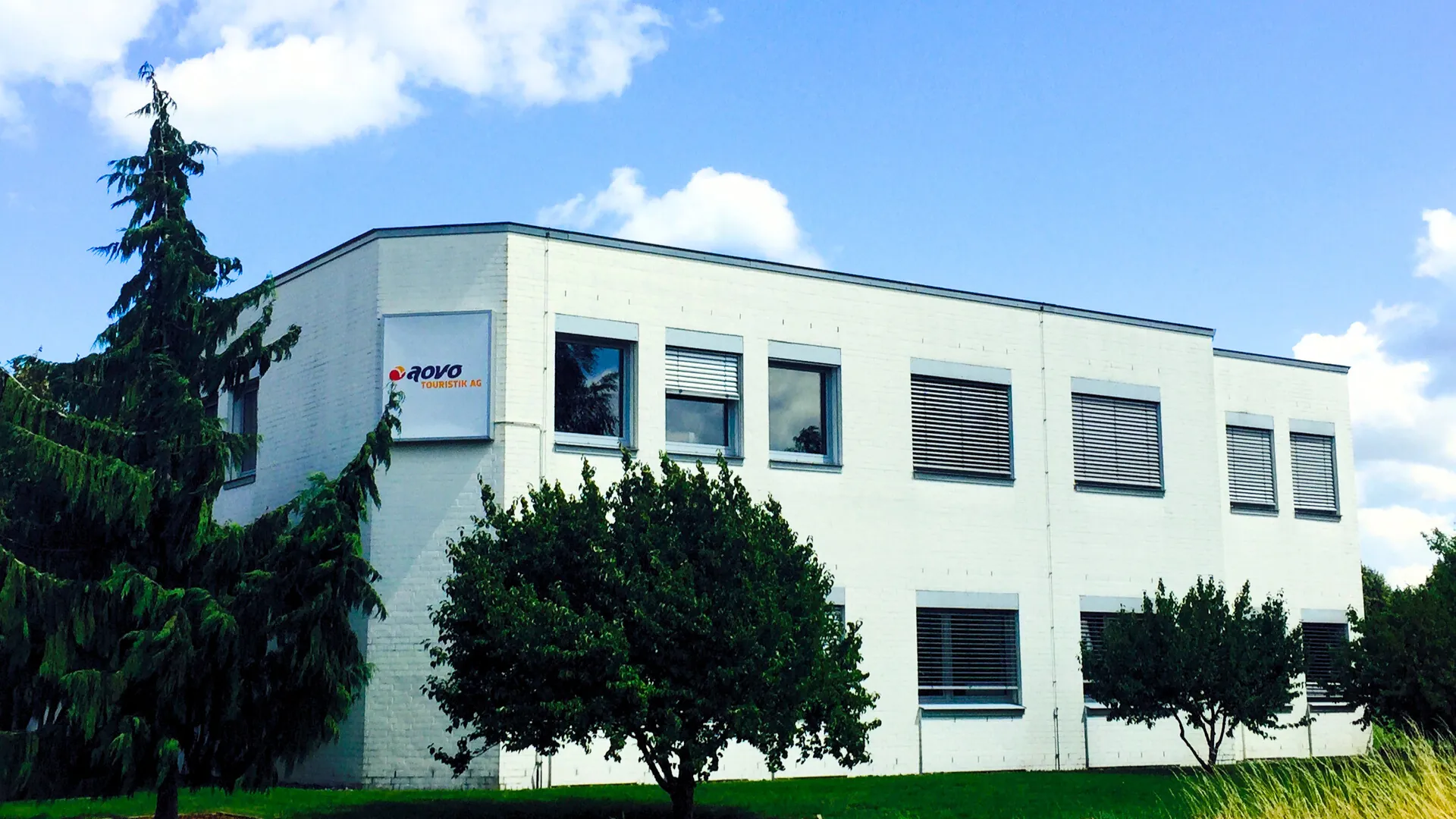 Außenansicht der aovo Touristik AG. Ein weißes zweistöckiges Gebäude, umgeben von grünen Pflanzen vor blauem Himmel mit kleinen Schleierwolken.