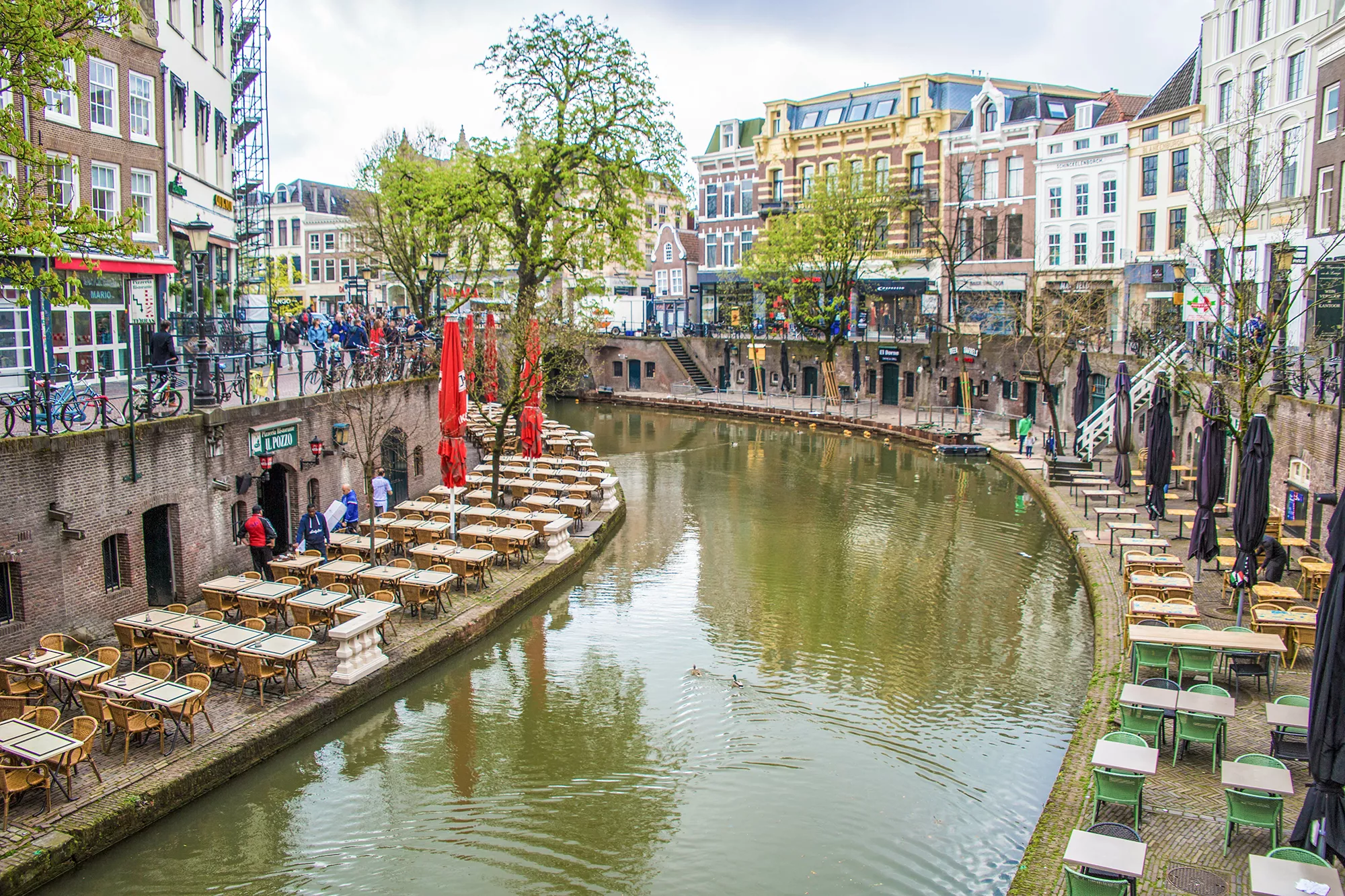 Das Bild zeigt eine idyllische Kanalszene in einer historischen Stadt. Entlang des Wassers befinden sich mehrere Terrassen mit leeren Tischen und Stühlen, die auf den Kanal ausgerichtet sind, sowie leuchtend rote Sonnenschirme. Die Gebäude entlang des Kanals sind mehrstöckig und haben klassische Fassaden. Auf der oberen Ebene sind Fußgänger zu sehen, während auf der unteren Ebene das ruhige Wasser des Kanals fließt. Bäume und vereinzelte Dekorationen verleihen der Szene eine gemütliche und einladende Atmosphäre.
