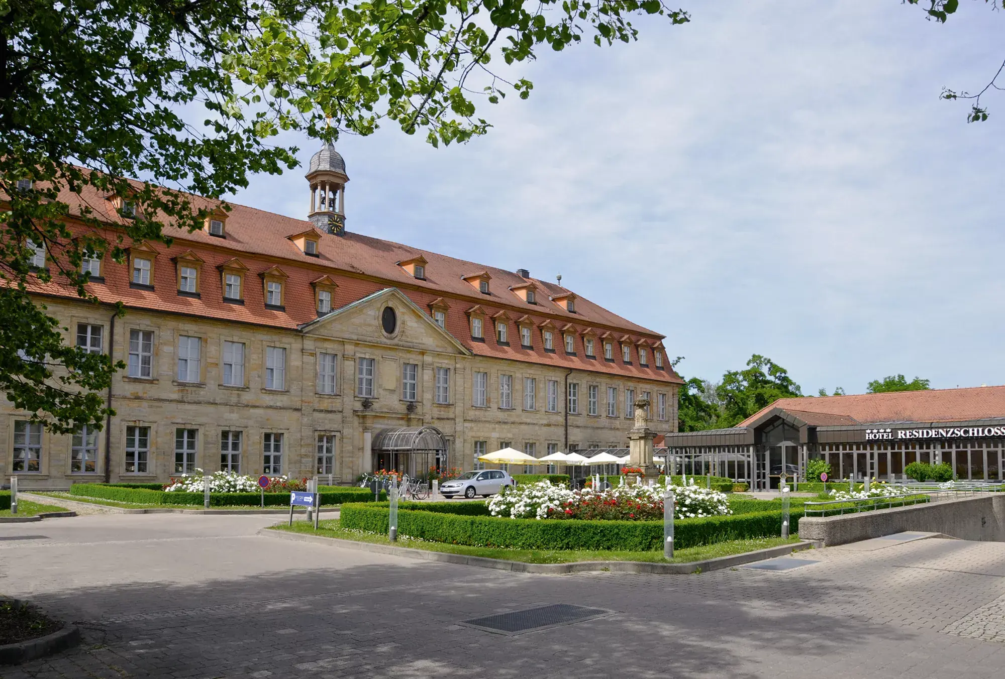 Schloss mit rotem Dach und Glockenturm, davor gepflegter Garten und Eingangsbereich des Welcome Hotel Residenzschloss Bamberg