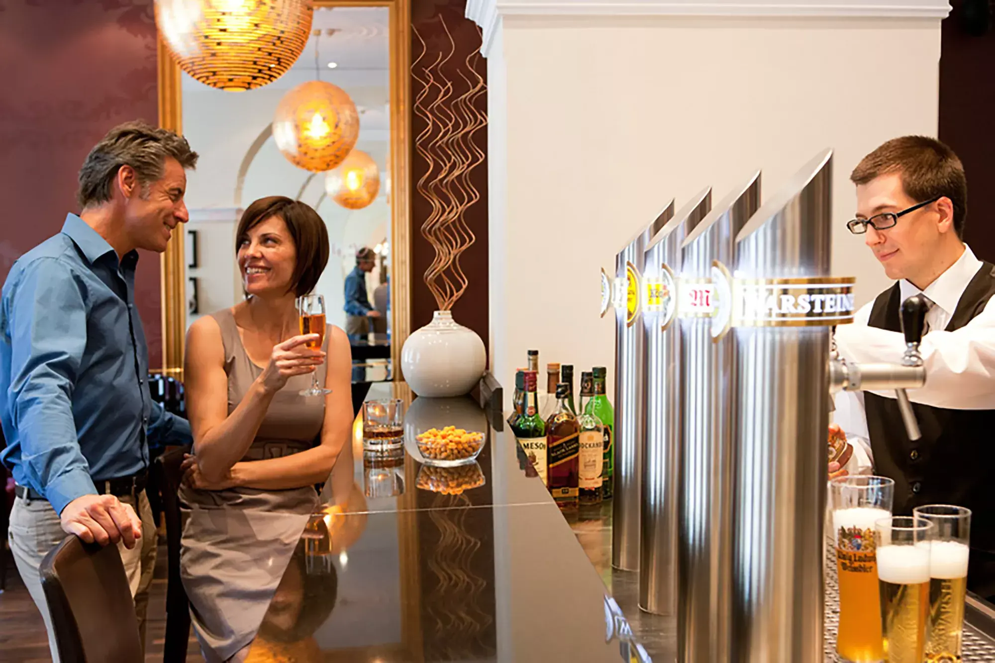 Zwei Gäste lehnen an einer Bar und unterhalten sich, während ein Barkeeper hinter der Theke Bier zapft.