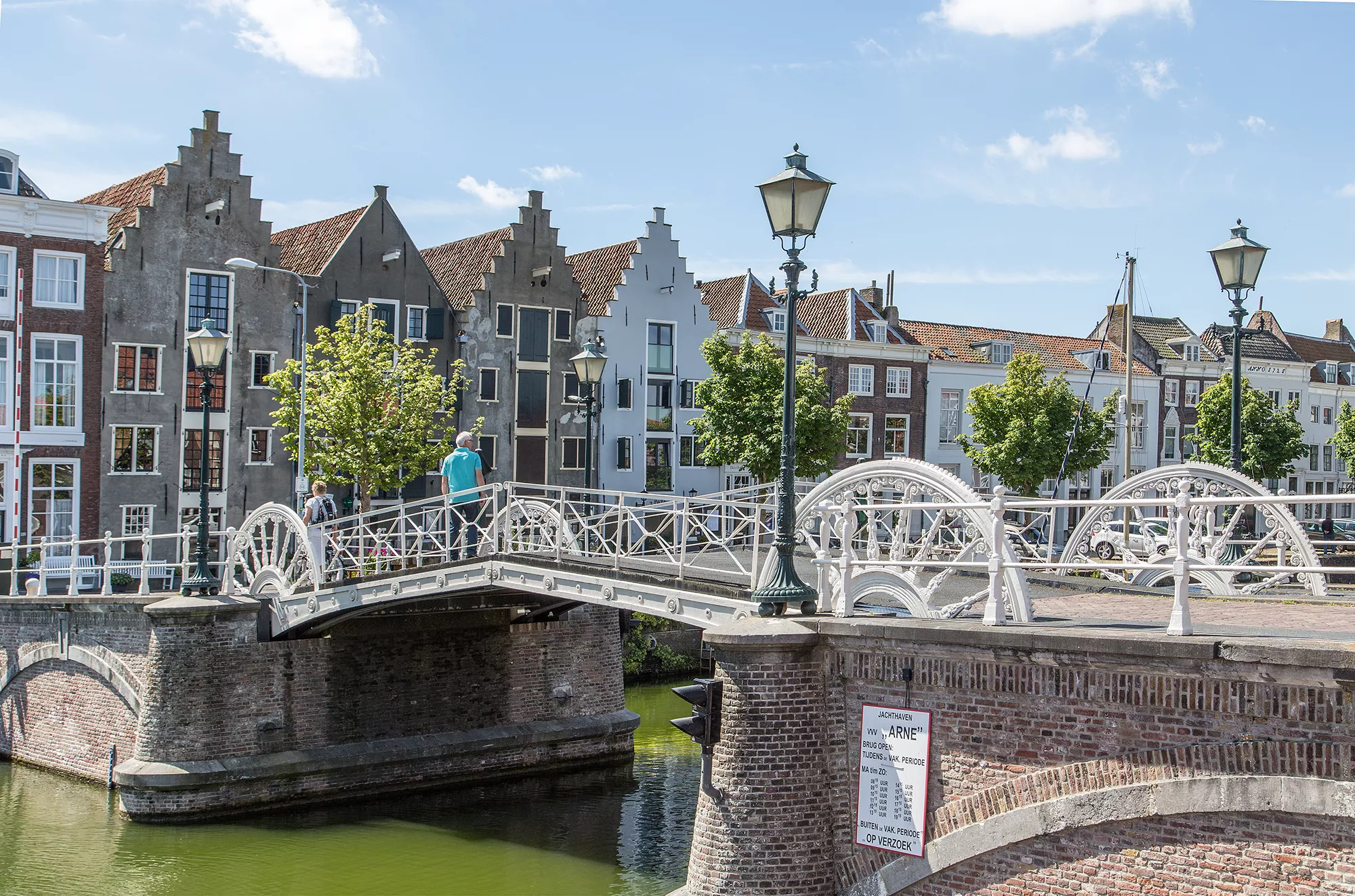 Das Bild zeigt die Spijkerbrug in Zeeland, eine malerische Brücke mit eleganten weißen Geländern und dekorativen Details, die sich über einen ruhigen Kanal spannt. Im Hintergrund stehen charmante Stadthäuser mit typisch niederländischen Giebeln, deren Fassaden in verschiedenen Farben gehalten sind. Auf der Brücke gehen einige Passanten spazieren, während die Laternen entlang des Geländers eine nostalgische Atmosphäre schaffen. Die Szene vermittelt das Flair einer historischen Stadt in Zeeland, eingebettet in eine ruhige und idyllische Umgebung.