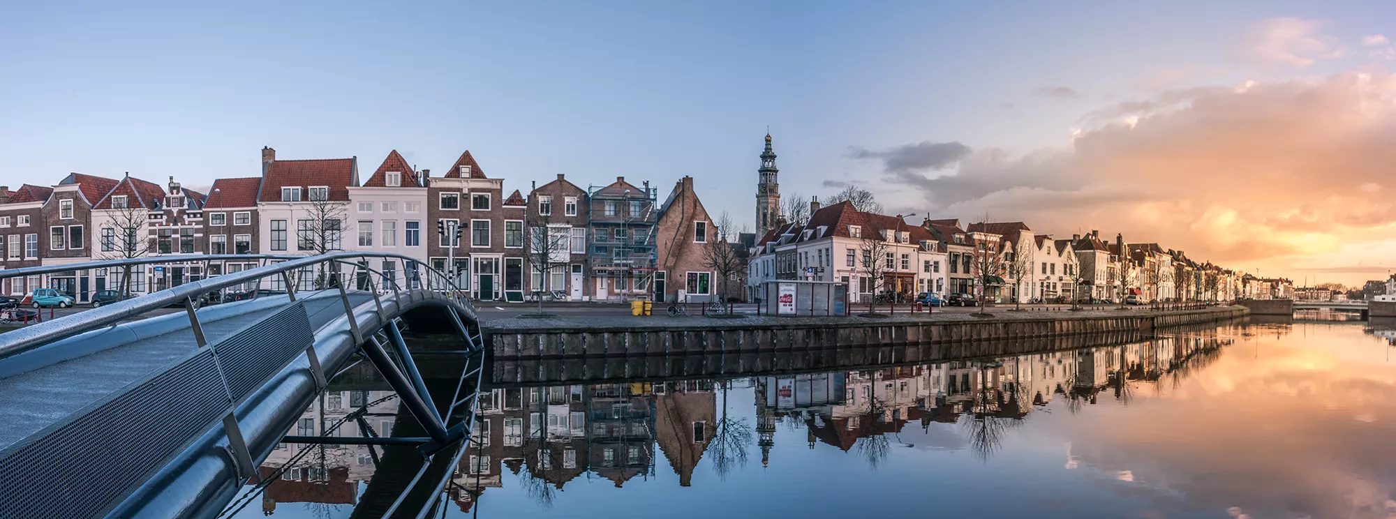 Das Bild zeigt eine Reihe von traditionellen Häusern entlang eines Kanals in Zeeland. Die Gebäude spiegeln sich klar im ruhigen Wasser, was eine harmonische und ruhige Atmosphäre schafft. Eine moderne Brücke führt über den Kanal und verbindet die beiden Ufer. Im Hintergrund erhebt sich ein Kirchturm, der das historische Ambiente der Umgebung unterstreicht. Der Himmel ist in sanften Farben des Sonnenuntergangs getaucht, was das Bild in ein warmes, weiches Licht hüllt und die idyllische Szenerie betont.