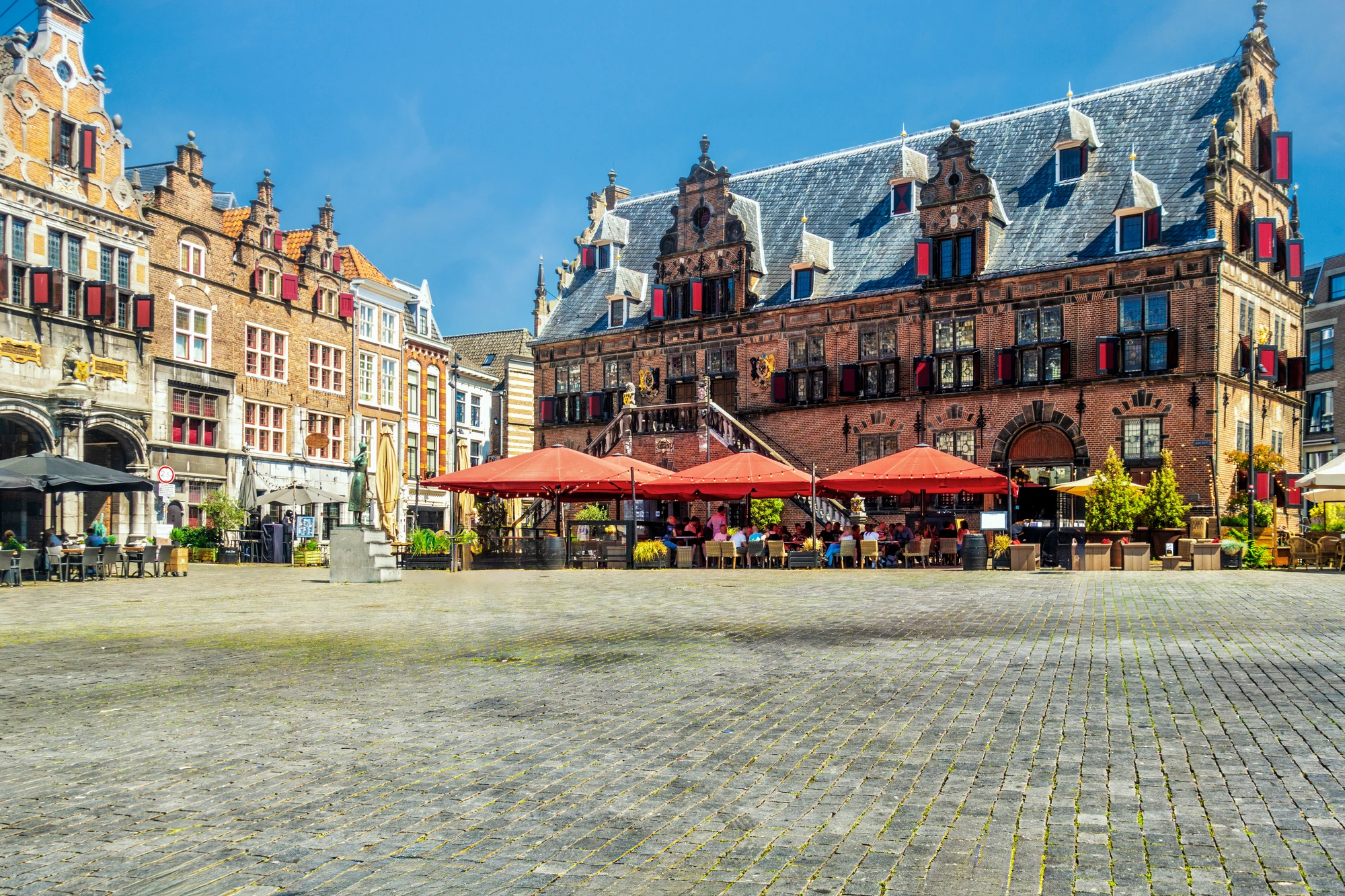 Der Grote Markt in Nijmegen an einem sonnigen Tag, mit entspannten Besuchern, die auf der Außenterrasse eines historischen Gebäudes sitzen. Das warme Sonnenlicht bringt die architektonischen Details des Gebäudes zur Geltung, während die lebhafte, aber gemütliche Atmosphäre des Platzes zum Verweilen einlädt.