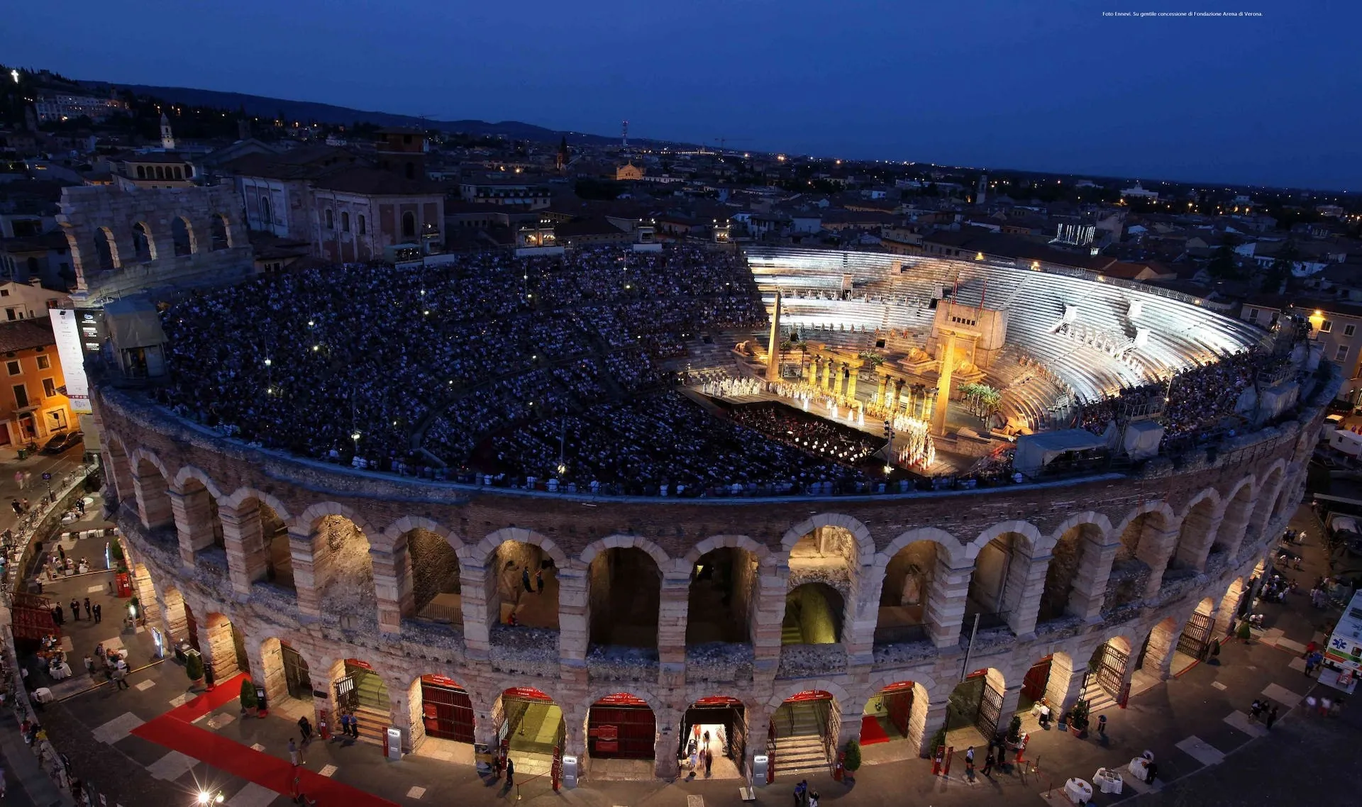 Die Arena di Verona ist in dieser Aufnahme während einer abendlichen Opernaufführung zu sehen. Die antiken Bögen des römischen Amphitheaters sind beleuchtet, während die Bühne mit einem imposanten Bühnenbild erstrahlt. Die Sitzreihen sind gut gefüllt, und das Publikum genießt die Vorstellung unter dem offenen Nachthimmel. Die historische Atmosphäre und das goldene Licht verleihen der Aufführung eine besondere Magie.