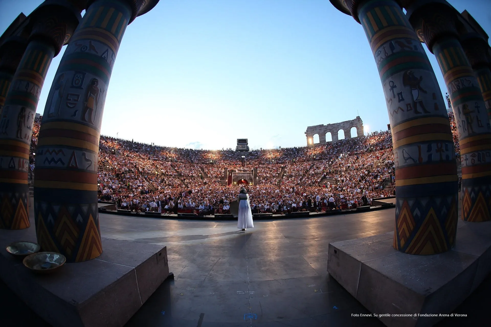 Die Bühne der Arena di Verona ist in dieser Aufnahme während einer Opernaufführung zu sehen. Majestätische, ägyptisch anmutende Säulen rahmen die Szene ein. Eine Sängerin steht im Mittelpunkt der Bühne und blickt auf das vollbesetzte Amphitheater, das von den antiken Bögen der Arena umgeben ist. Die Menschenmengen füllen die Ränge bis in die oberen Bereiche und schaffen eine beeindruckende Kulisse für das historische Opernerlebnis unter freiem Himmel.