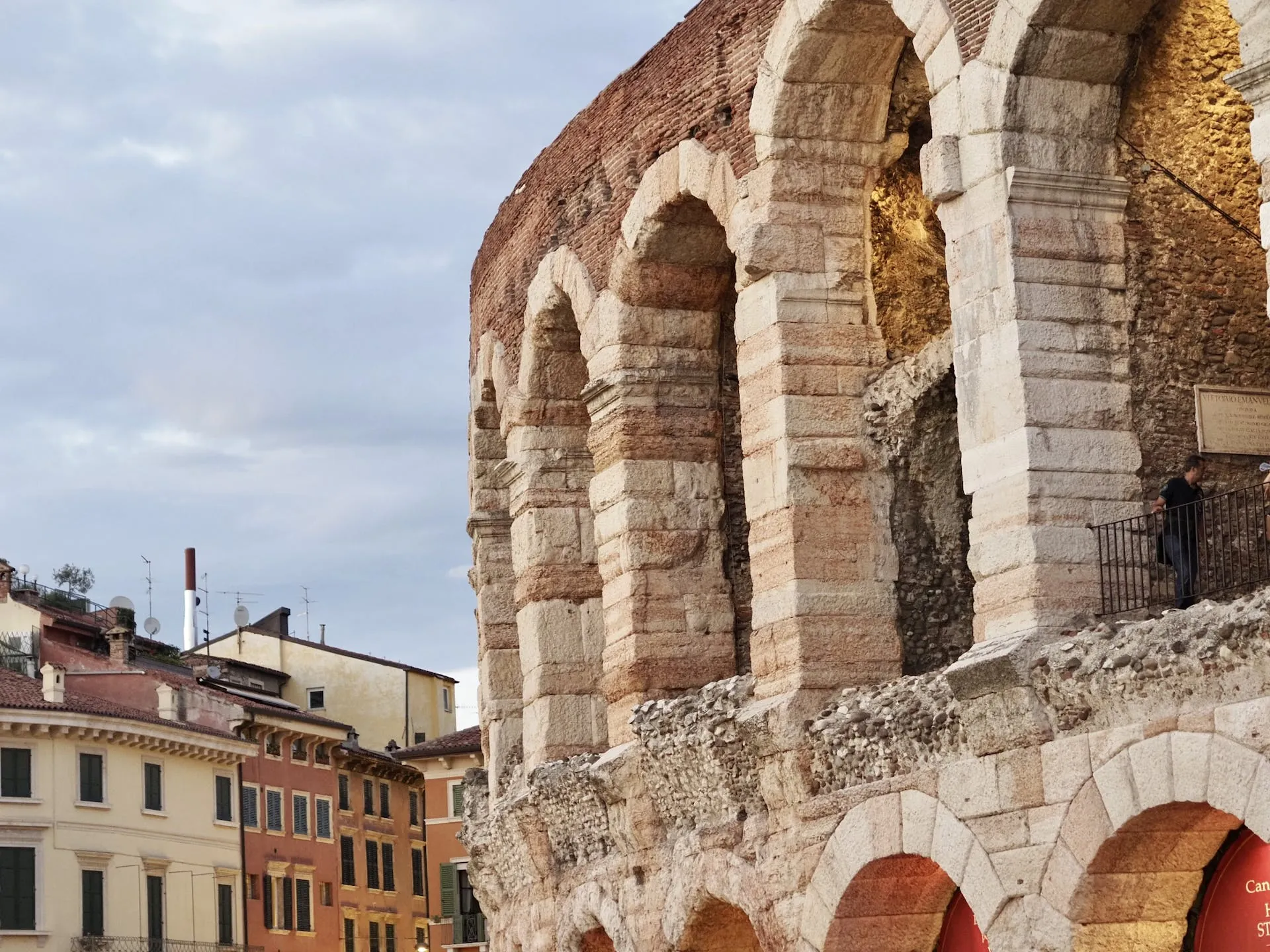 Die Fassade der Arena di Verona zeigt eindrucksvolle römische Architektur mit hohen Rundbögen und massivem Steinmauerwerk. Die antiken Steine haben durch die Jahrhunderte eine warme Patina angenommen, und die Struktur ist trotz ihres Alters gut erhalten. Im Hintergrund sind farbenfrohe italienische Gebäude zu sehen, die das historische Ambiente dieser berühmten Sehenswürdigkeit ergänzen.