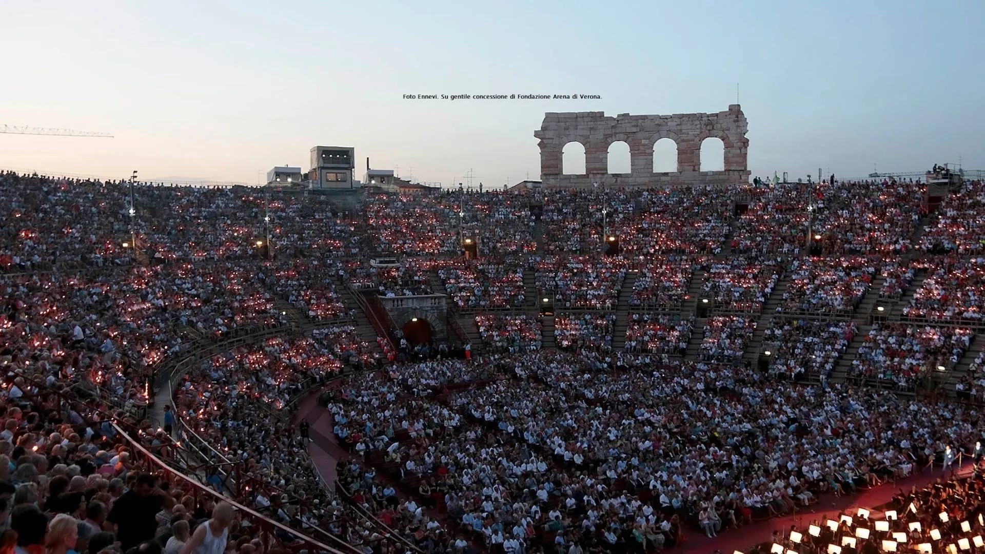 Das Bild zeigt das Publikum in der Arena di Verona während einer abendlichen Veranstaltung. Die Ränge des antiken Amphitheaters sind dicht gefüllt, und das Publikum hält unzählige kleine Kerzen in den Händen, was eine magische Atmosphäre im Halbdunkel schafft. Im Hintergrund ragen die antiken Mauern der Arena in den Abendhimmel, während die Spannung auf die bevorstehende Aufführung wächst. Die beeindruckende Kulisse und die Menge der Besucher unterstreichen die Bedeutung dieser historischen Spielstätte für spektakuläre Opernaufführungen.