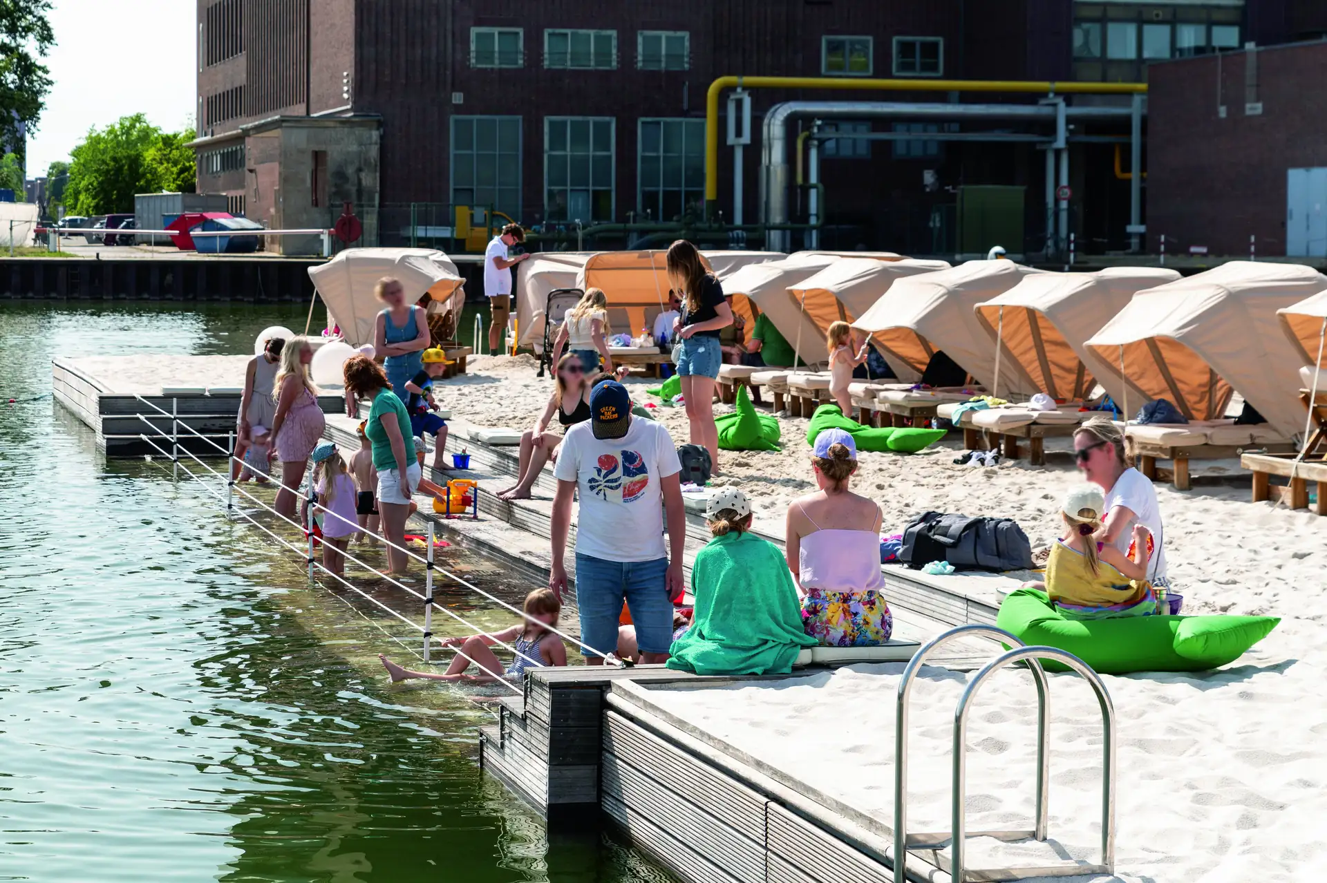 Autostadt Wolfsburg im Sommer 2024: Menschen entspannen und spielen am städtischen Sandstrand am Wasser mit Liegen und Sonnenschirmen im Hintergrund