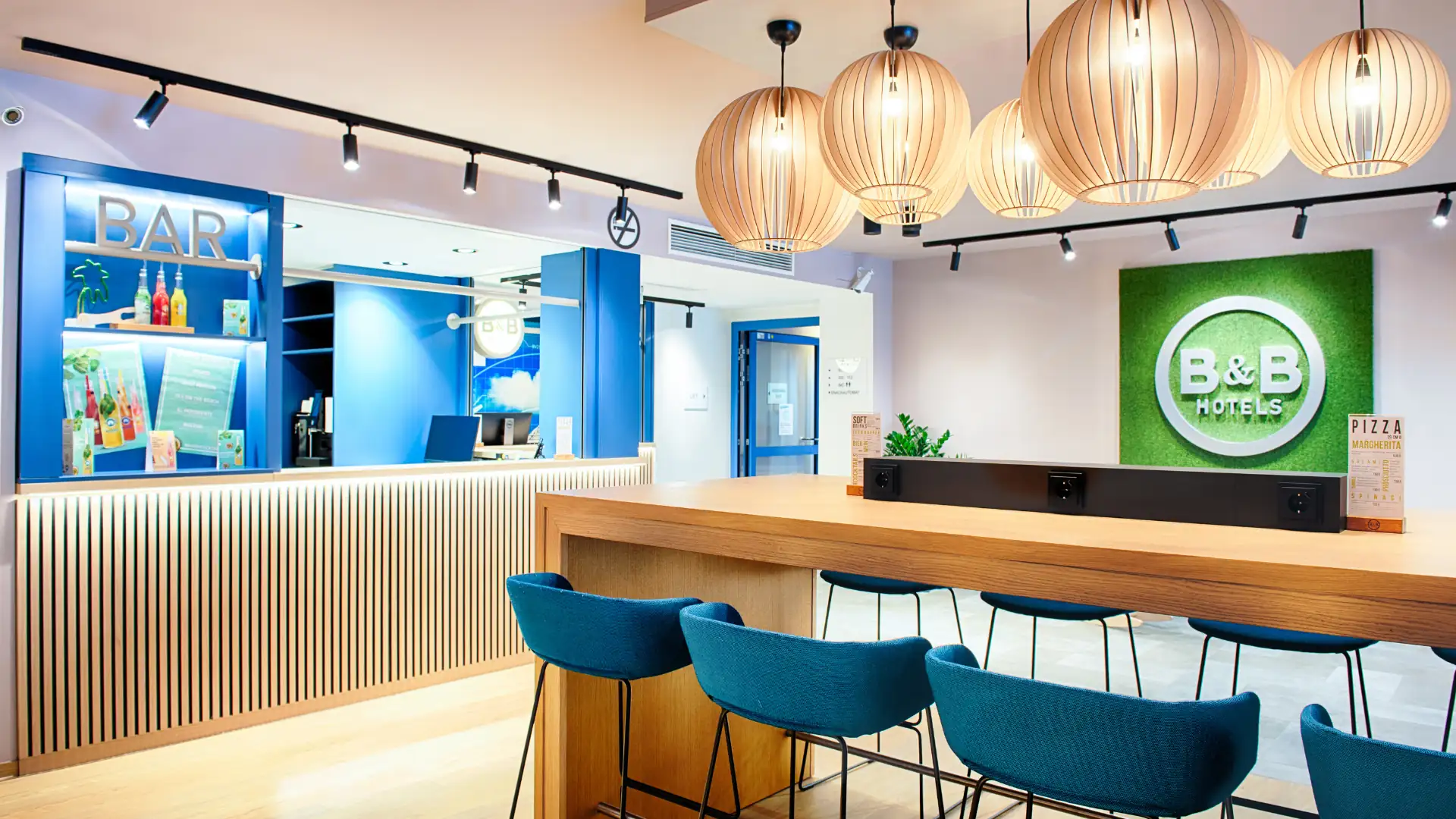 B&B Hotels im Kachelformat: Empfangsbereich des B&B Hotels München Airport mit blauem Barbereich, Holztresen und vier blauen Barhockern unter runden Deckenlampen.