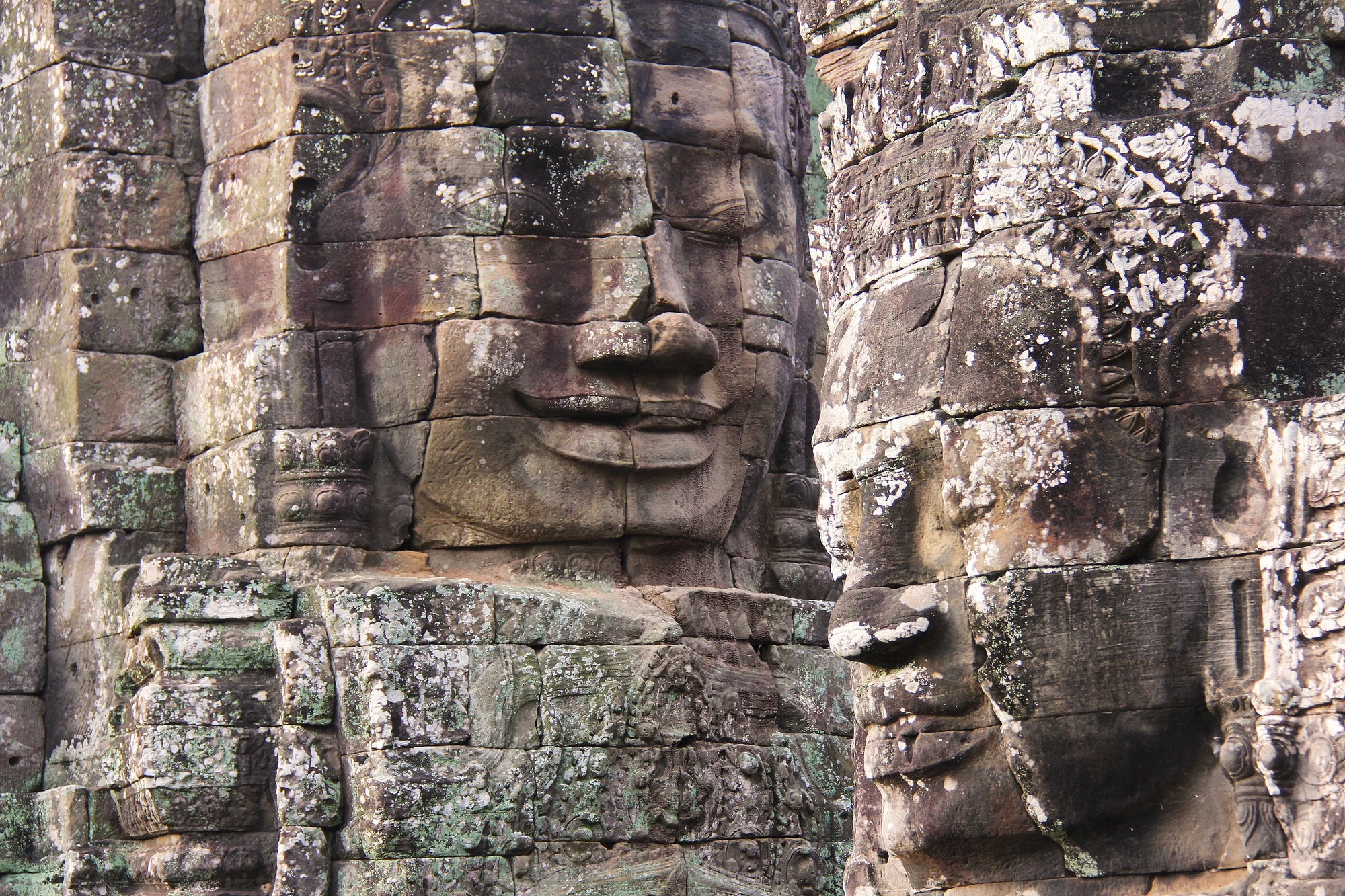 Zwei große Steingesichter des Bayon-Tempels in Angkor, mit detaillierten Gesichtszügen und Moosbewuchs auf den Steinen.