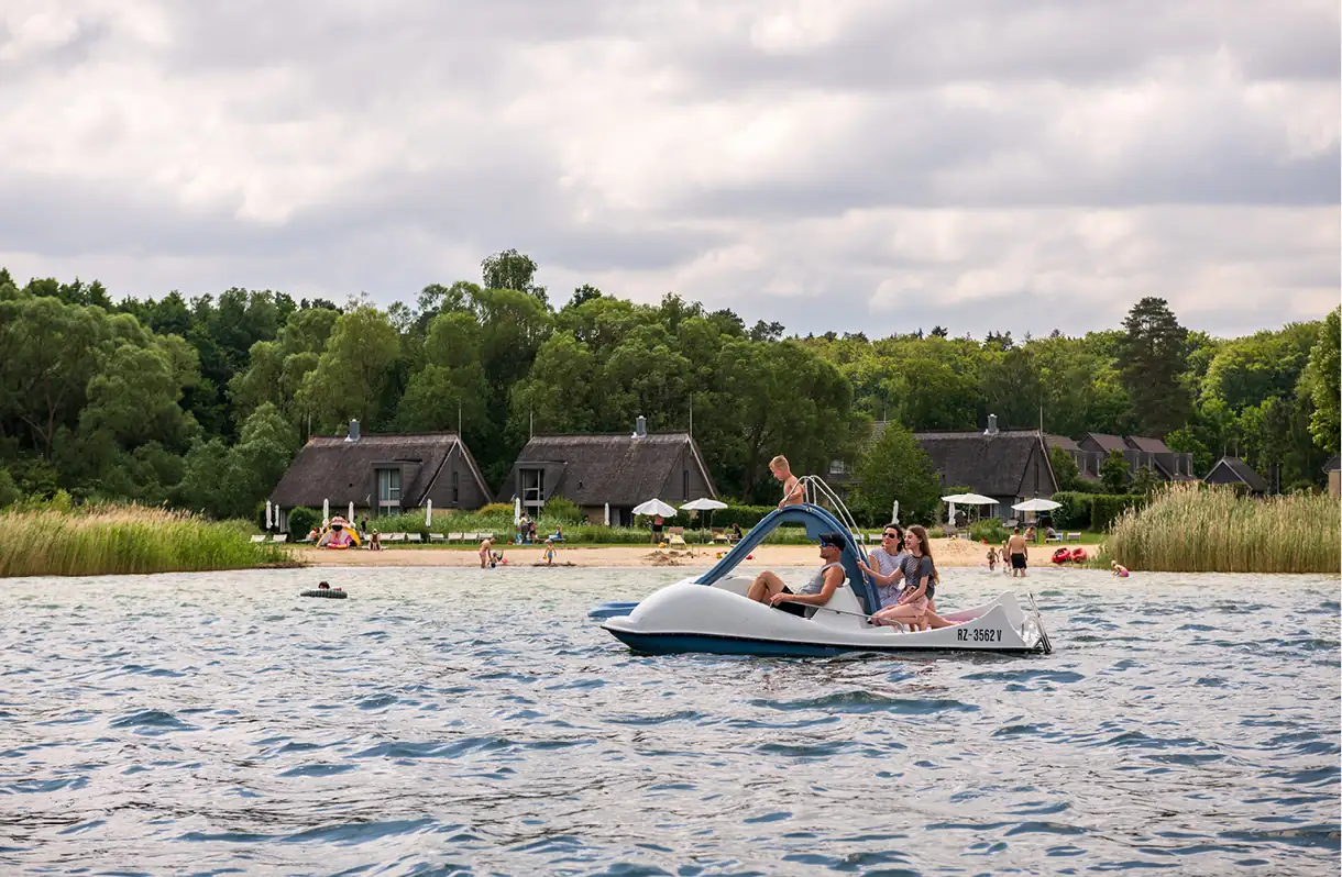 Beech Resort Plauer See: Menschen auf einem Tretboot auf dem Plauer See, im Hintergrund Häuser und Bäume unter bewölktem Himmel.