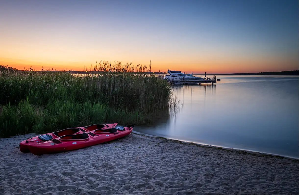 Beech Resort Plauer See: Rotes Doppelkajak am Sandufer des Plauer Sees bei Sonnenuntergang, im Hintergrund Steg mit Booten und Schilf.