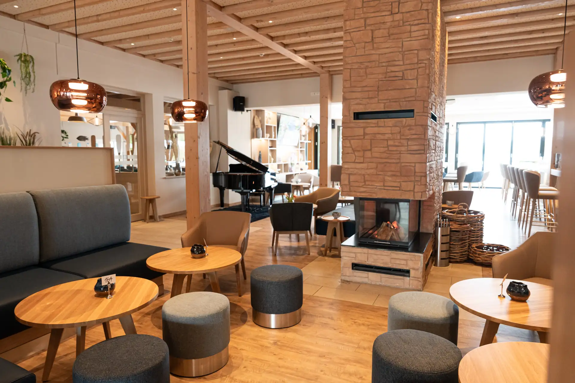 Beech Resort Plauer See: Gemütlicher Lobby mit runden Holztischen, grauen Hockern, Kamin mit Steinverkleidung und Klavier im Hintergrund.