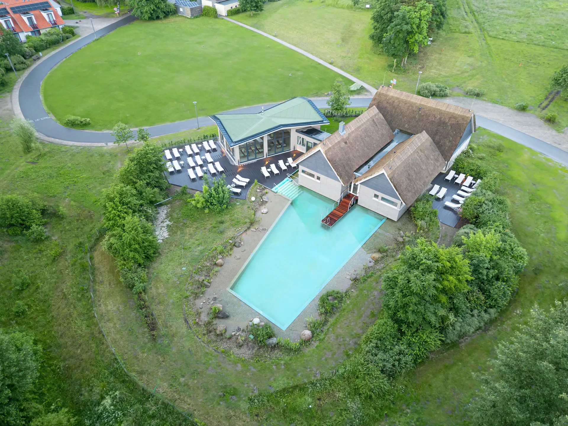 Beech Resort Plauer See: Luftaufnahme des Wald SPA mit Reetdach, angrenzendem Schwimmbecken und Liegestühlen auf einer Terrasse, umgeben von Grünflächen und Wegen.