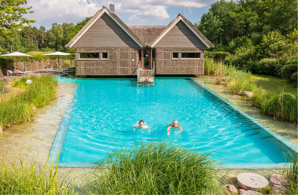 Beech Resort Plauer See: Zwei Personen schwimmen in einem rechteckigen Pool vor einem Holzhaus mit zwei Giebeln, umgeben von Grün und Bäumen.