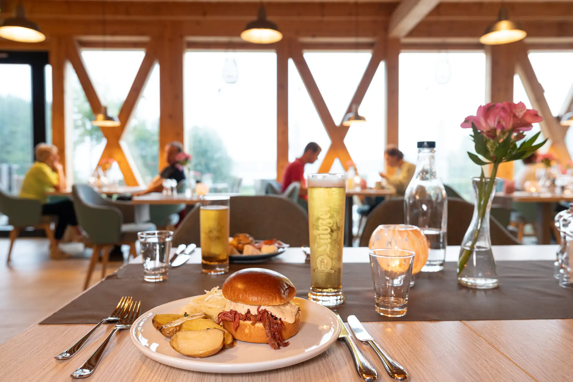 Beech Resort Plauer See: Tisch mit Burger, Pommes, Getränken und Besteck im Restaurant mit Holzbalken und Gästen im Hintergrund.