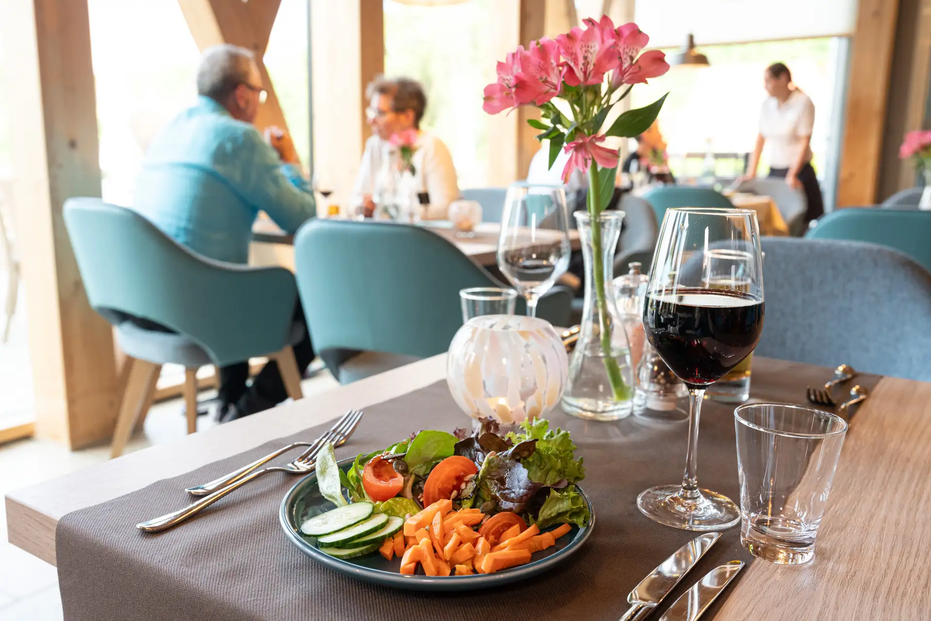 Beech Resort Plauer See: Teller mit gemischtem Salat aus Tomaten, Gurken, Karotten und Blattsalat auf gedecktem Tisch mit Weinglas, Wasserglas und Blumenvasen in Restaurant.