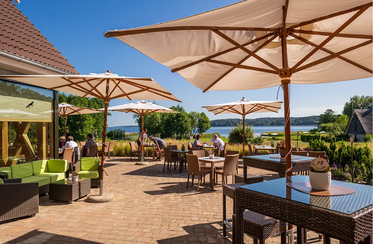 Beech Resort Plauer See: Terrassenbereich mit Sonnenschirmen, Sitzgruppen aus Korbgeflecht und Menschen, im Hintergrund See und Bäume.