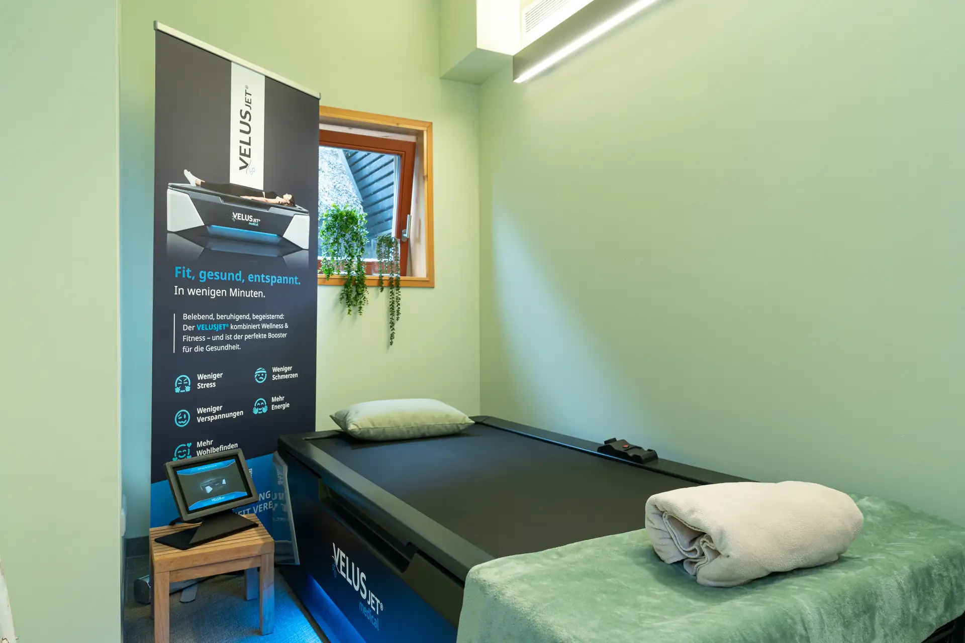 Beech Resort Plauer See: Behandlungsraum mit VELUSjet-Massagegerät, Liege mit Kissen und Handtuch, Tablet auf kleinem Holztisch, Fenster mit Pflanzen.