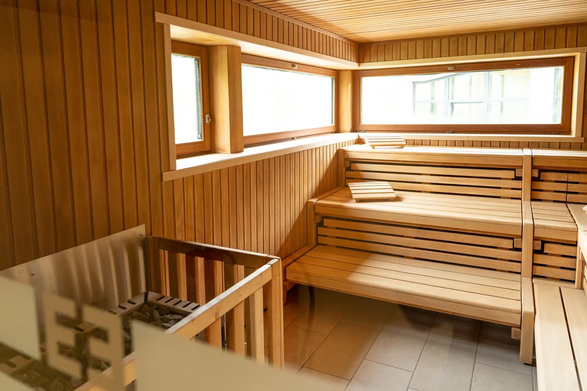 Beech Resort Plauer See: Saunaraum mit Holzbänken in zwei Ebenen, Holzverkleidung an Wänden, großen Fenstern und Saunaofen mit Steinen.