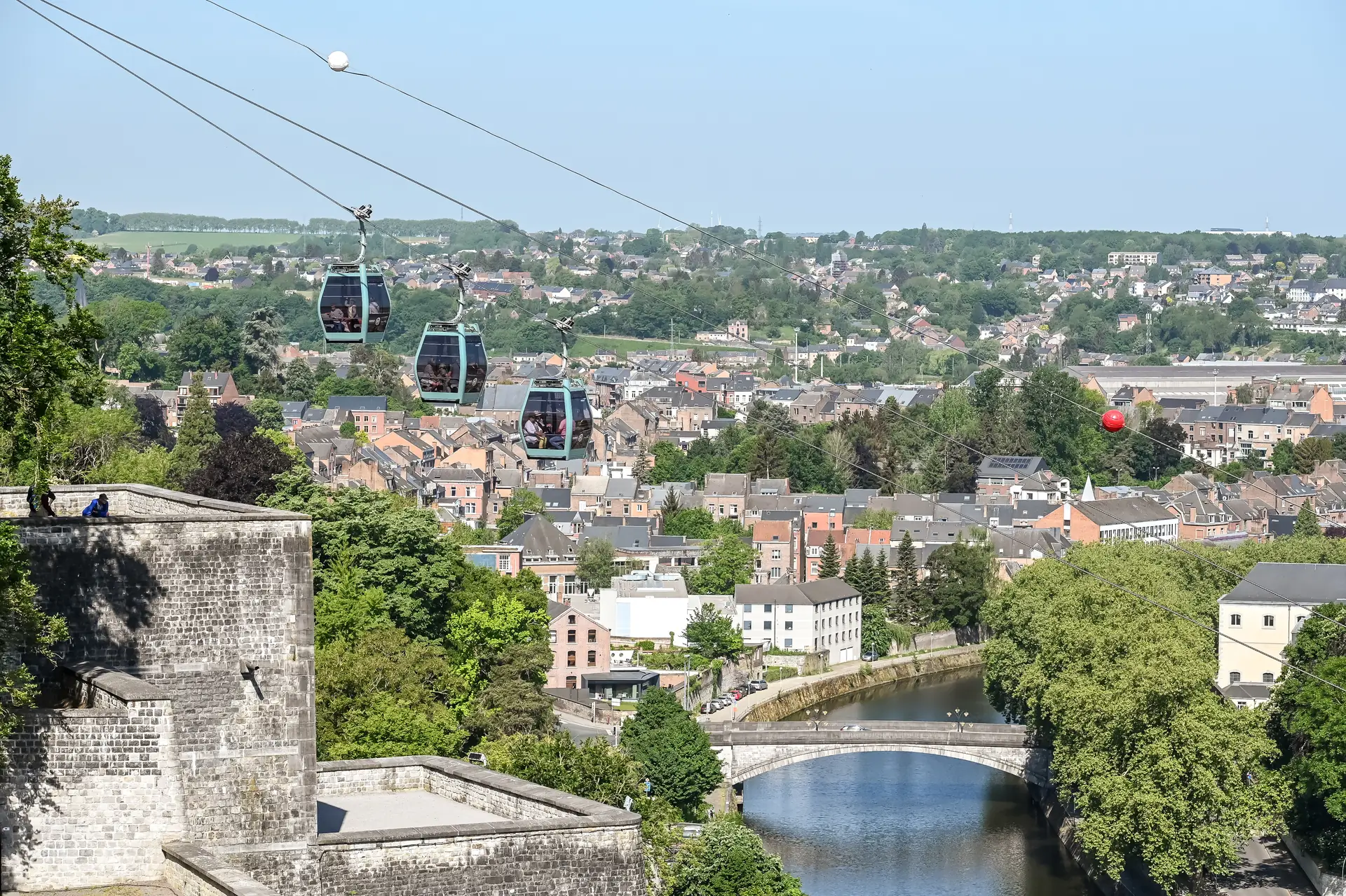 Drei Kabinen der neuen Seilbahn von Namur in der Luft, mit Blick auf die Dächer der historischen Altstadt und die umliegende Landschaft bei Tageslicht, die eine beeindruckende Perspektive auf die Stadt bietet.