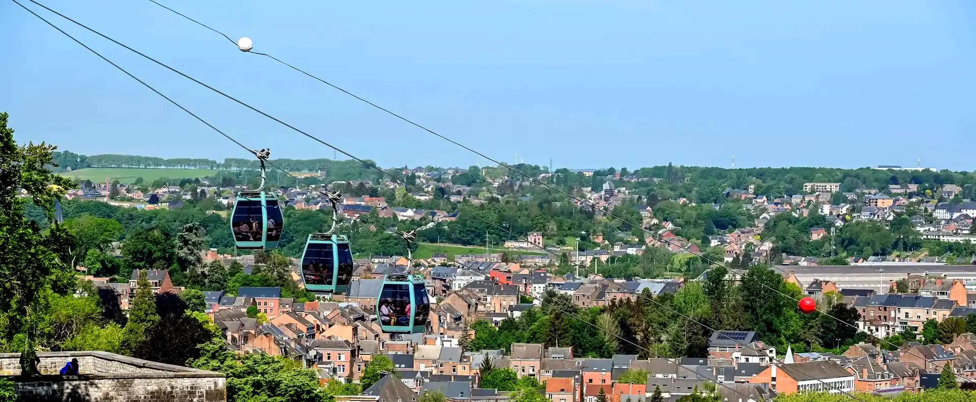 Drei Kabinen der neuen Seilbahn von Namur in der Luft, mit Blick auf die Dächer der historischen Altstadt und die umliegende Landschaft bei Tageslicht, die eine beeindruckende Perspektive auf die Stadt bietet.