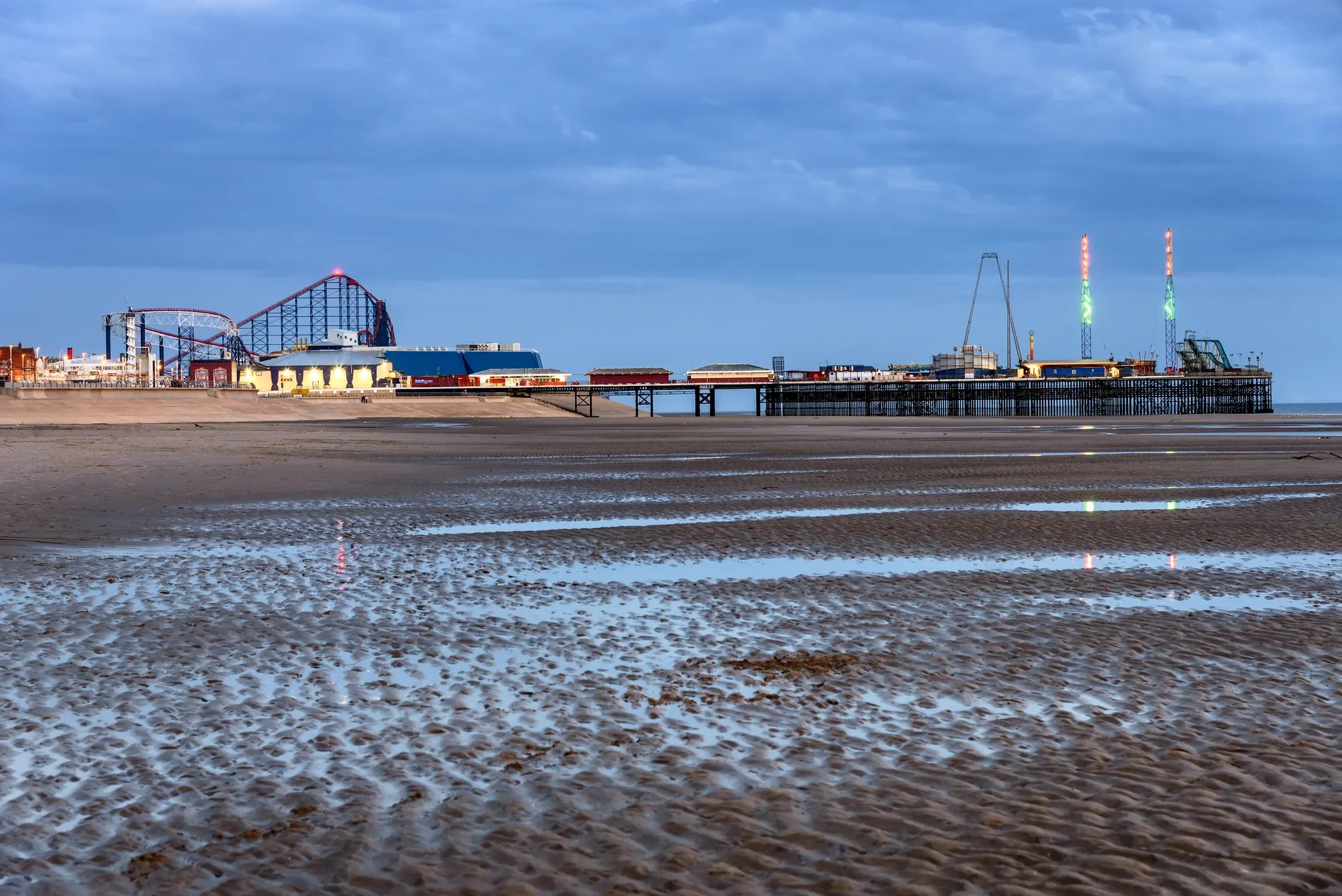 Das Bild zeigt den Blackpool Pleasure Beach in Großbritannien, aufgenommen bei Ebbe mit einem weiten Blick über den sandigen Strand. Im Hintergrund sind die markanten Achterbahnen und Fahrgeschäfte des Freizeitparks sichtbar, während die hell erleuchtete Promenade eine lebendige Atmosphäre andeutet. Der blaue Himmel und die ruhige Wasseroberfläche, die sich in Pfützen auf dem Strand sammelt, verleihen der Szene eine besondere Abendstimmung und betonen die Verbindung zwischen der Küste und den spannenden Attraktionen des Parks.