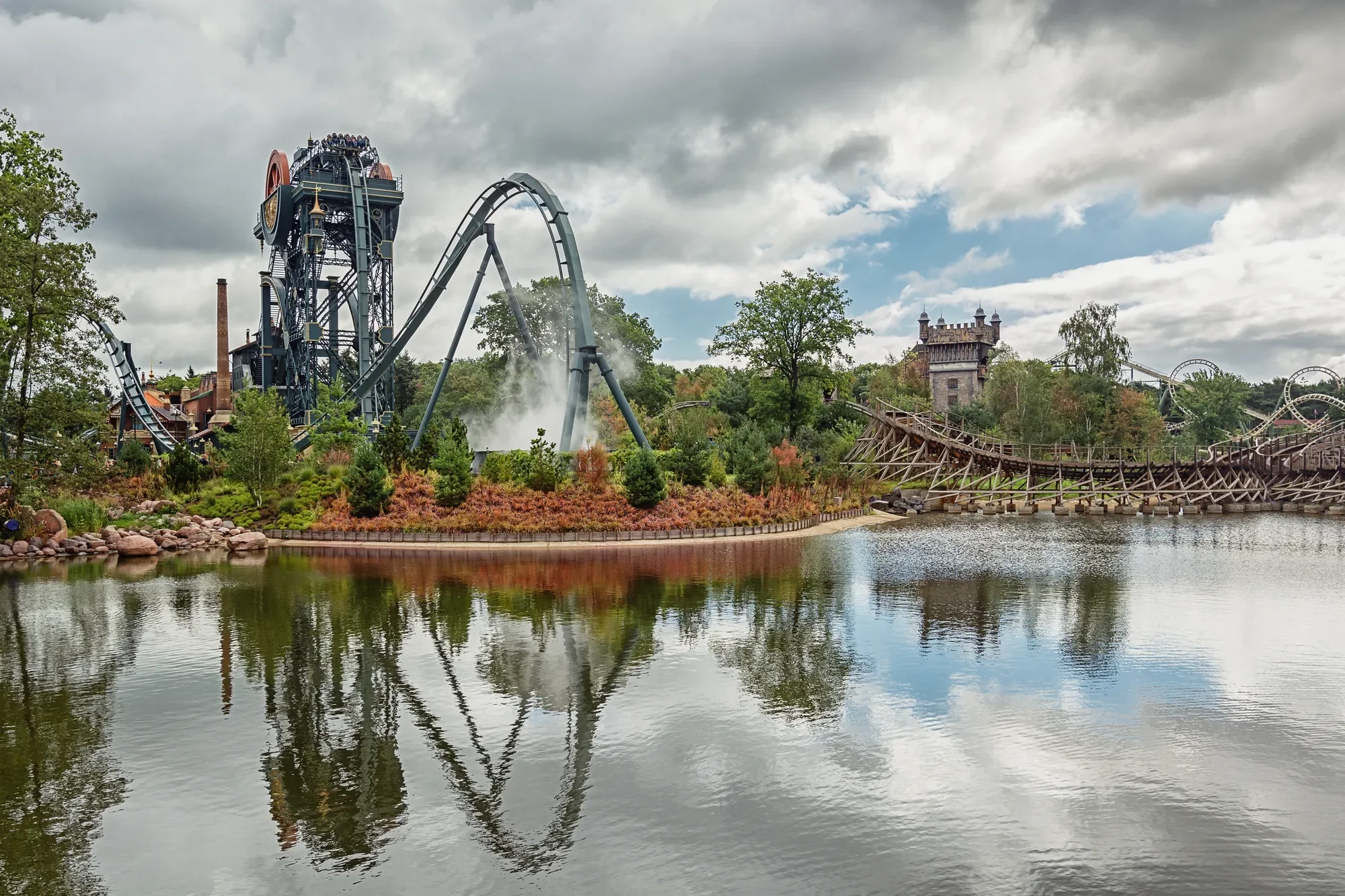Das Bild zeigt den Freizeitpark Efteling in den Niederlanden, mit einer eindrucksvollen Achterbahn, die sich imposant in die Landschaft einfügt. Die reflektierenden Wasserflächen im Vordergrund und die umgebenden Bäume schaffen eine harmonische Atmosphäre, die im Kontrast zur dynamischen Stahlkonstruktion der Achterbahn steht. Die detailreiche Gestaltung des Parks und die Verbindung von Natur und Attraktion machen Efteling zu einem einzigartigen Erlebnis für Besucher.