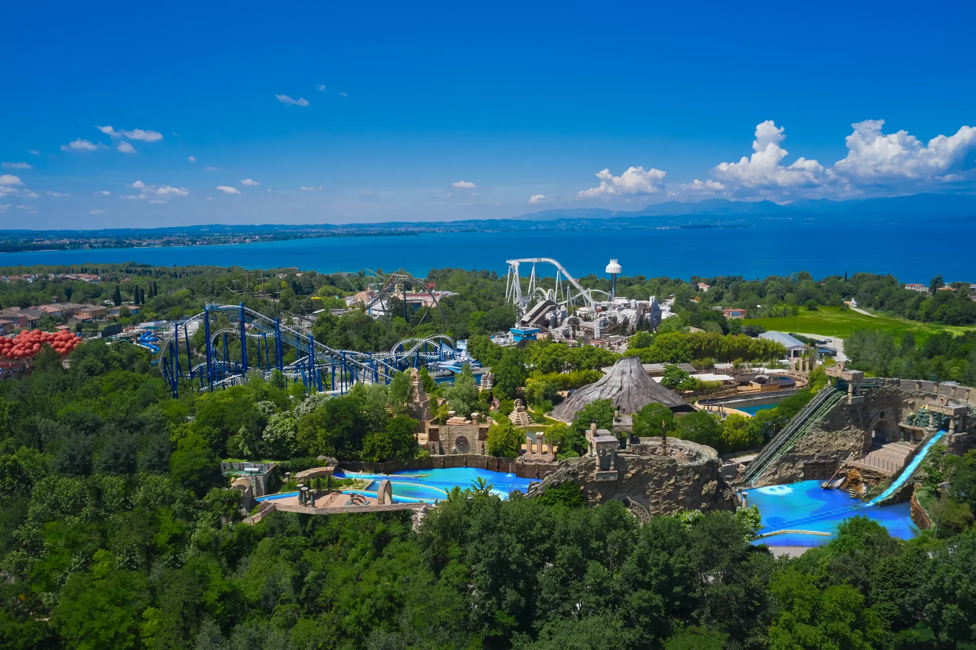 Das Bild zeigt eine Panoramaaufnahme des Gardaland Freizeitparks in Italien, eingebettet in üppiges Grün mit dem malerischen Gardasee im Hintergrund. Die verschiedenen Achterbahnen und Attraktionen schlängeln sich durch die üppige Landschaft, während helle Farben und Wasserfahrgeschäfte die aufregende und lebendige Atmosphäre des Parks betonen. Die Szenerie vermittelt eine Mischung aus Abenteuer, Vergnügen und landschaftlicher Schönheit, was Gardaland zu einem beliebten Ziel für Familien und Besucher aller Altersgruppen macht.