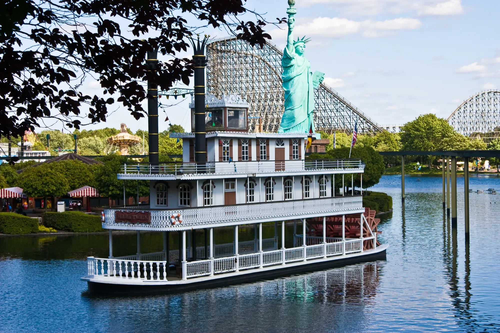 Das Bild zeigt den Heide Park Soltau mit einem markanten Raddampfer und der Nachbildung der Freiheitsstatue im Vordergrund. Im Hintergrund sind große Achterbahnen zu sehen, die das vielfältige Freizeitangebot des Parks verdeutlichen. Das ruhige Wasser des Sees spiegelt die Szenen wider und verleiht dem Bild eine besondere Atmosphäre, die Abenteuer und Entspannung vereint.