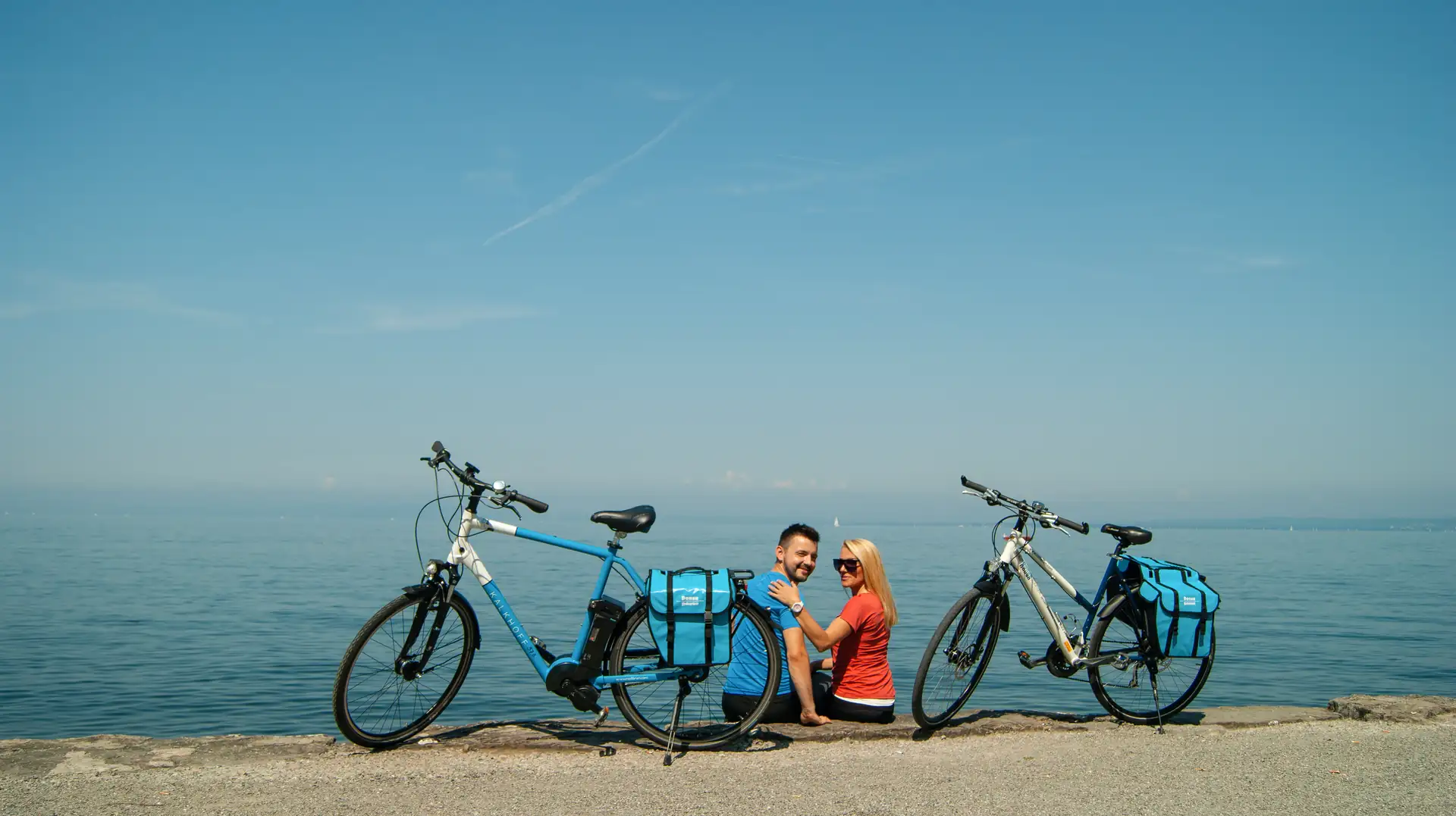 Zwei Personen sitzen am Wasser zwischen zwei Fahrrädern mit blauen Taschen, Blick auf den Bodensee