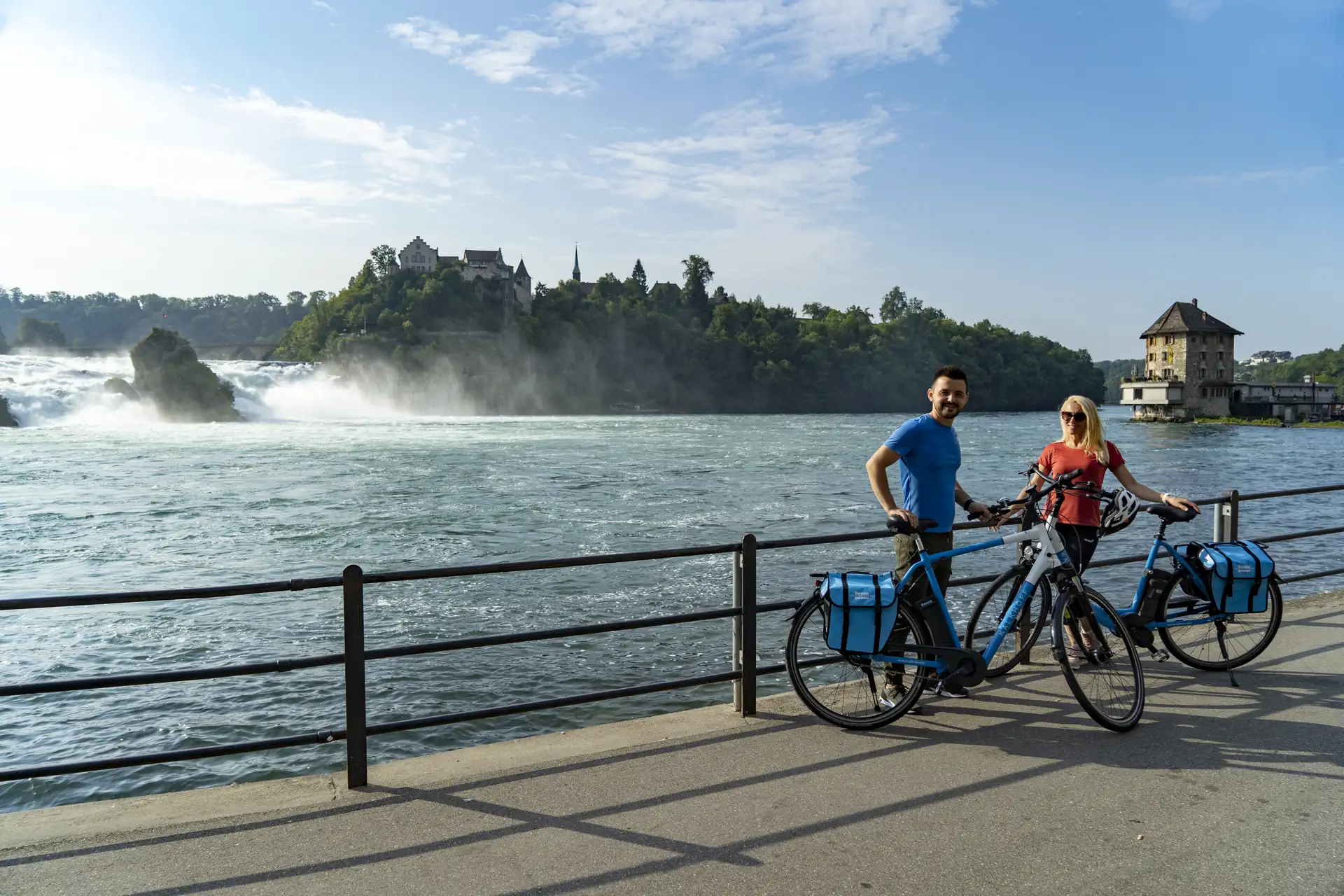 Zwei Radfahrer mit blauen Fahrradtaschen stehen am Rheinufer mit Blick auf den Rheinfall in Schaffhausen und eine Burg im Hintergrund.