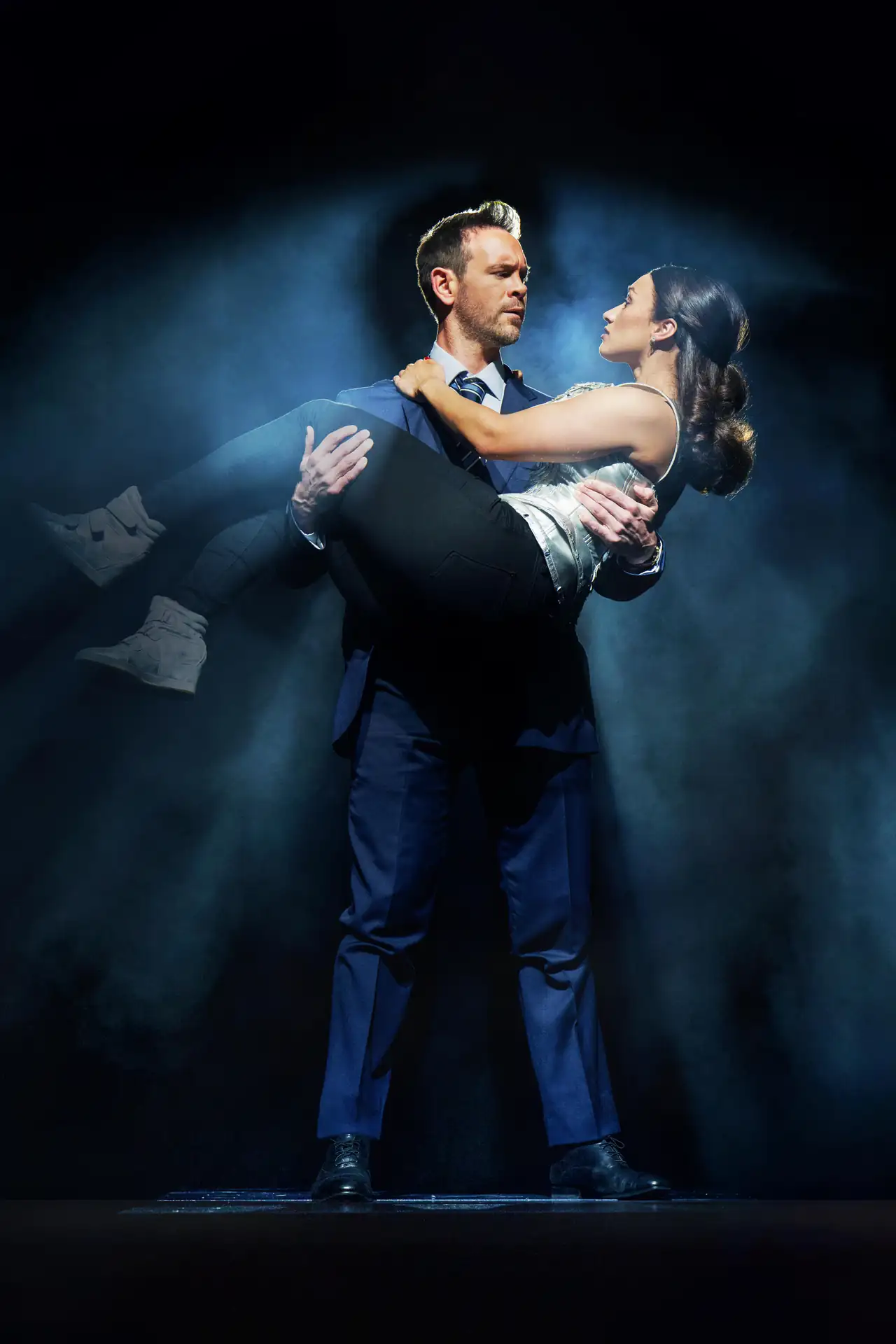 The Bodyguard – The Musical Tour: Ein Mann im blauen Anzug hält eine Frau in schwarzer Hose und weißem Oberteil im Arm, beide stehen auf einer dunklen Bühne mit Nebel.